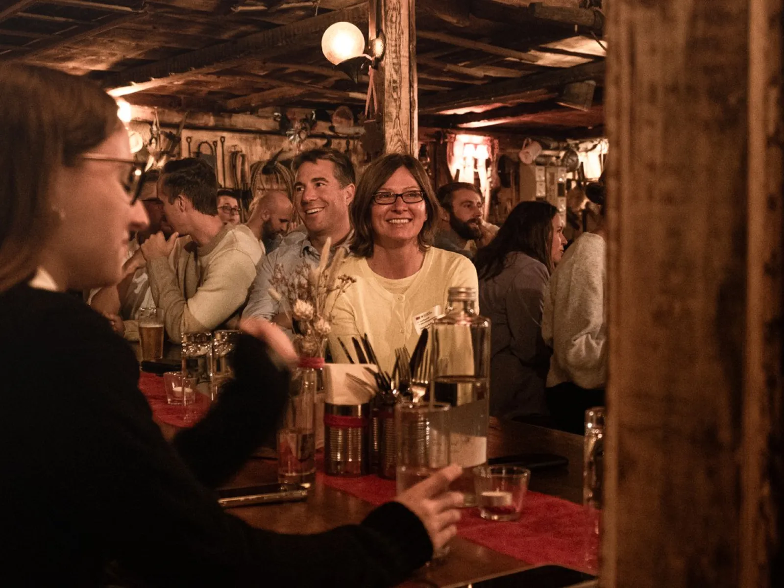 Eine Gruppe von Menschen sitzt in einer gemütlichen, rustikalen Bar. Im Vordergrund unterhalten sich Personen an einem Tisch, während im Hintergrund weitere Gäste lächeln und die Atmosphäre genießen. Die Szene wirkt lebhaft und einladend.