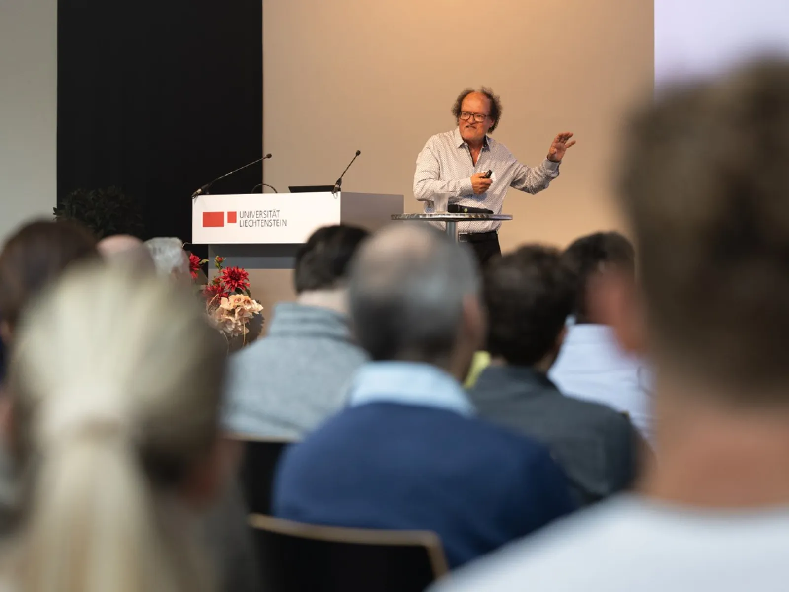 Ein Redner steht an einem Podium und spricht zu einem Publikum bei einer Veranstaltung an der Universität Liechtenstein. Im Hintergrund ist das Logo der Universität sichtbar.