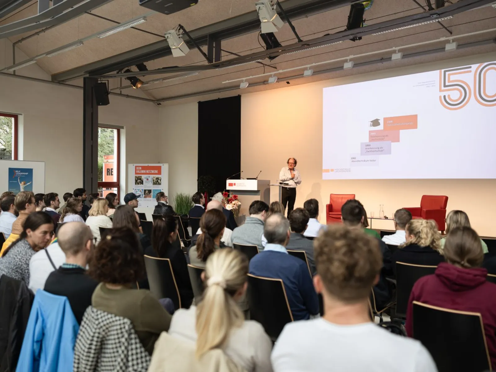 Ein Vortragender steht auf einer Bühne vor einem Publikum in einem modernen Veranstaltungsraum. Auf der Leinwand hinter ihm ist eine Präsentation mit der Zahl "50" zu sehen. Die Zuhörer sitzen aufmerksam in Reihen.