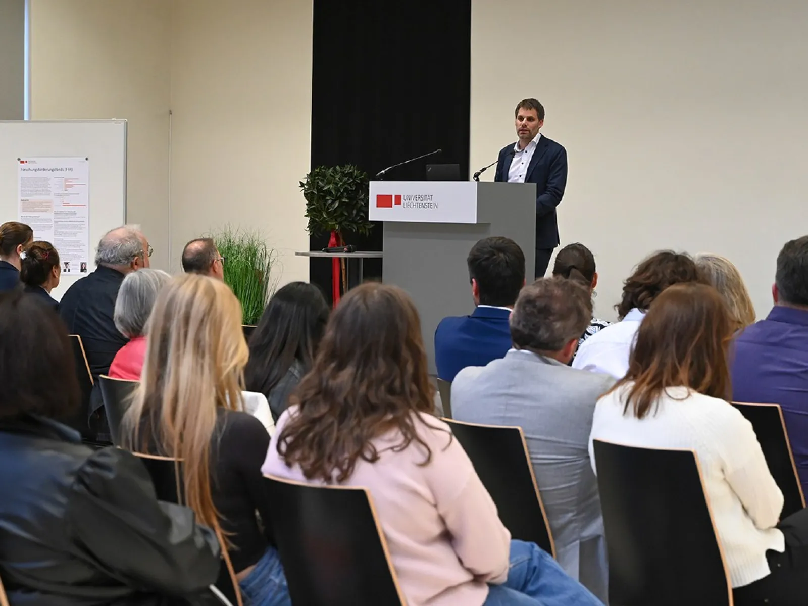 Rektor Christian Frommelt hält eine Rede am Rednerpult vor einem sitzenden Publikum in einem Konferenzraum. Im Hintergrund ist ein Poster sichtbar.