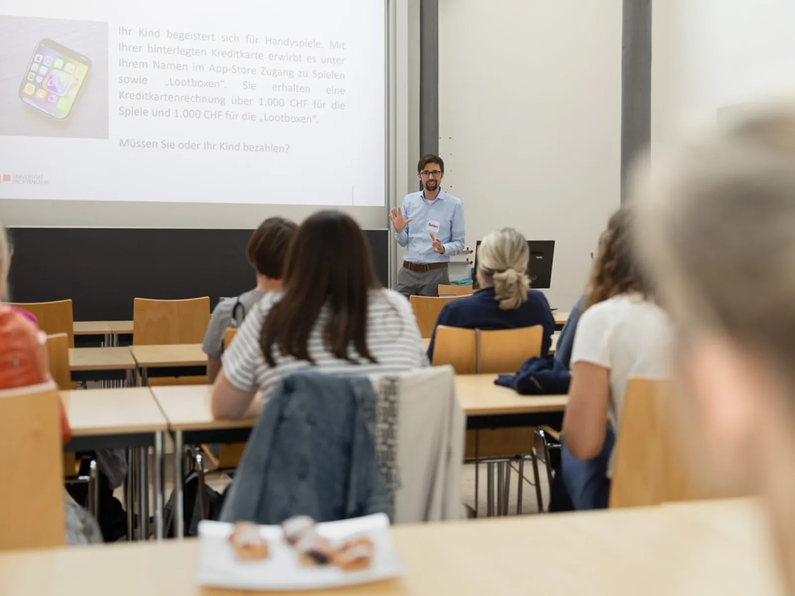 Ein Dozent hält einen Vortrag in einem Seminarraum vor mehreren Erwachsenen. Die Zuhörer sitzen an Tischen und schauen zur Präsentation an der Leinwand.