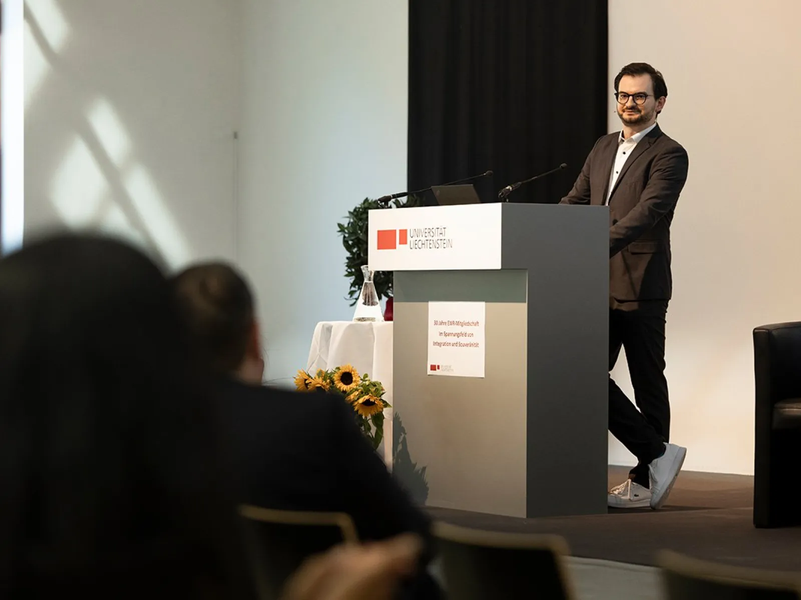 Ein Mann steht an einem Rednerpult in einem Konferenzraum der Universität Liechtenstein. Er trägt einen Anzug und spricht zu einem Publikum. Im Hintergrund sind Sonnenblumen und ein Fenster zu sehen.