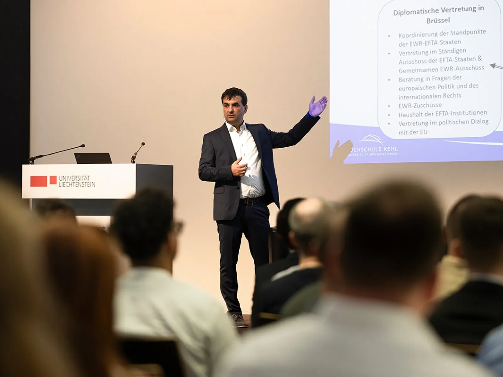 Ein Mann hält einen Vortrag vor einem Publikum an der Universität Liechtenstein. Er steht neben einer Leinwand mit einer Präsentation. Im Vordergrund sind die Köpfe der Zuhörer zu sehen.
