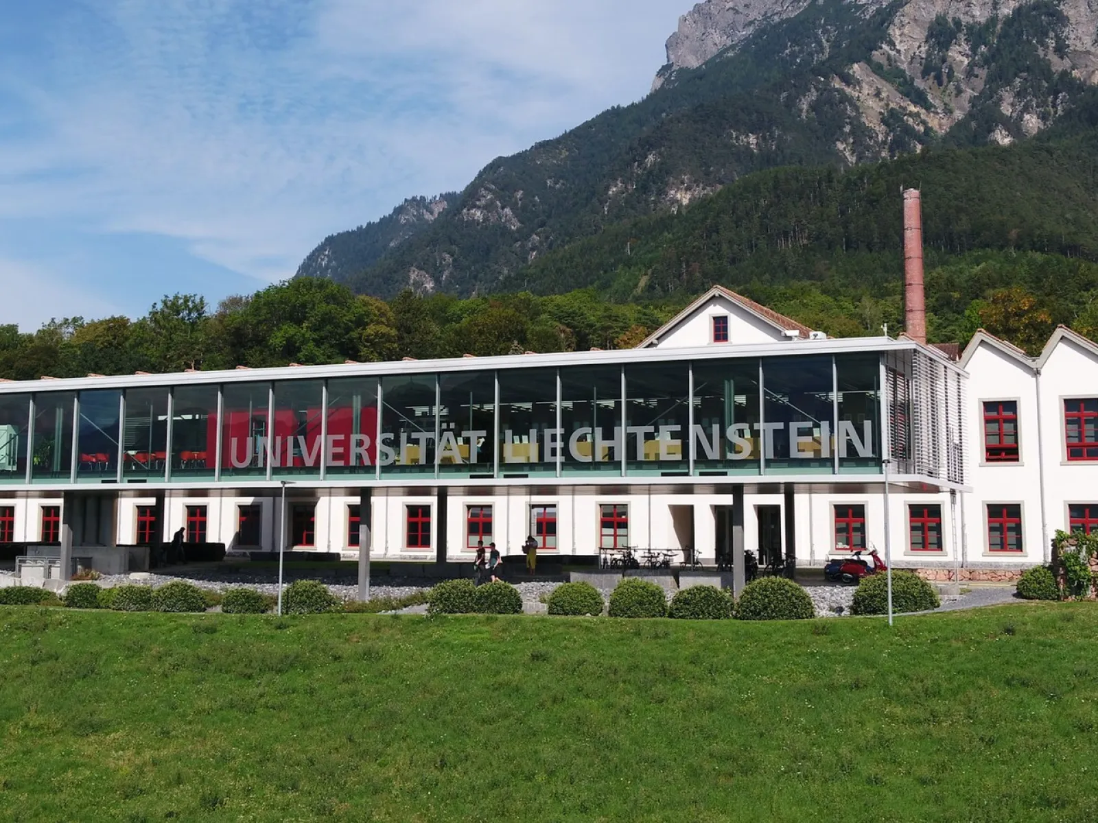 Außenansicht der Universität Liechtenstein mit modernem Glasgebäude und traditionellem weißen Gebäude im Hintergrund, umgeben von grüner Landschaft und Bergen.