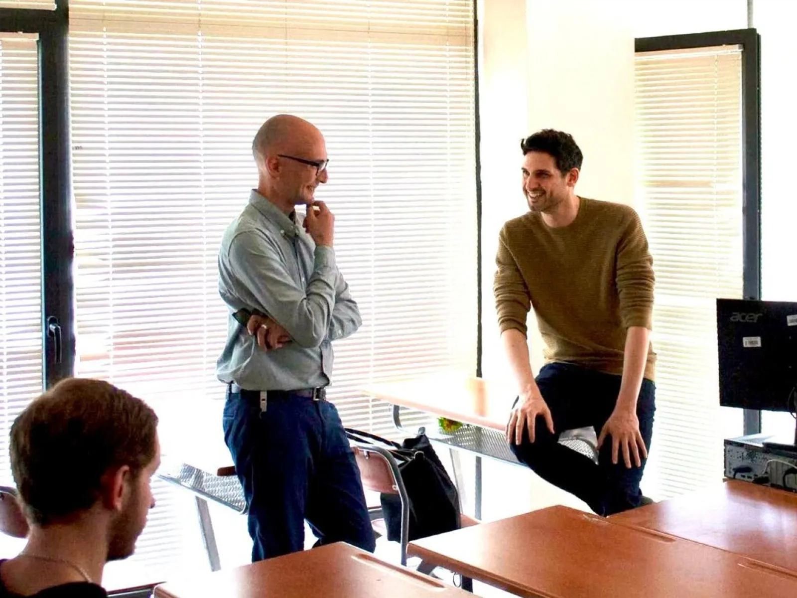 Zwei Männer in einem hellen Raum stehen und sitzen lächelnd an einem Tisch, in einem Gespräch vertieft. Im Hintergrund sind Jalousien zu sehen.