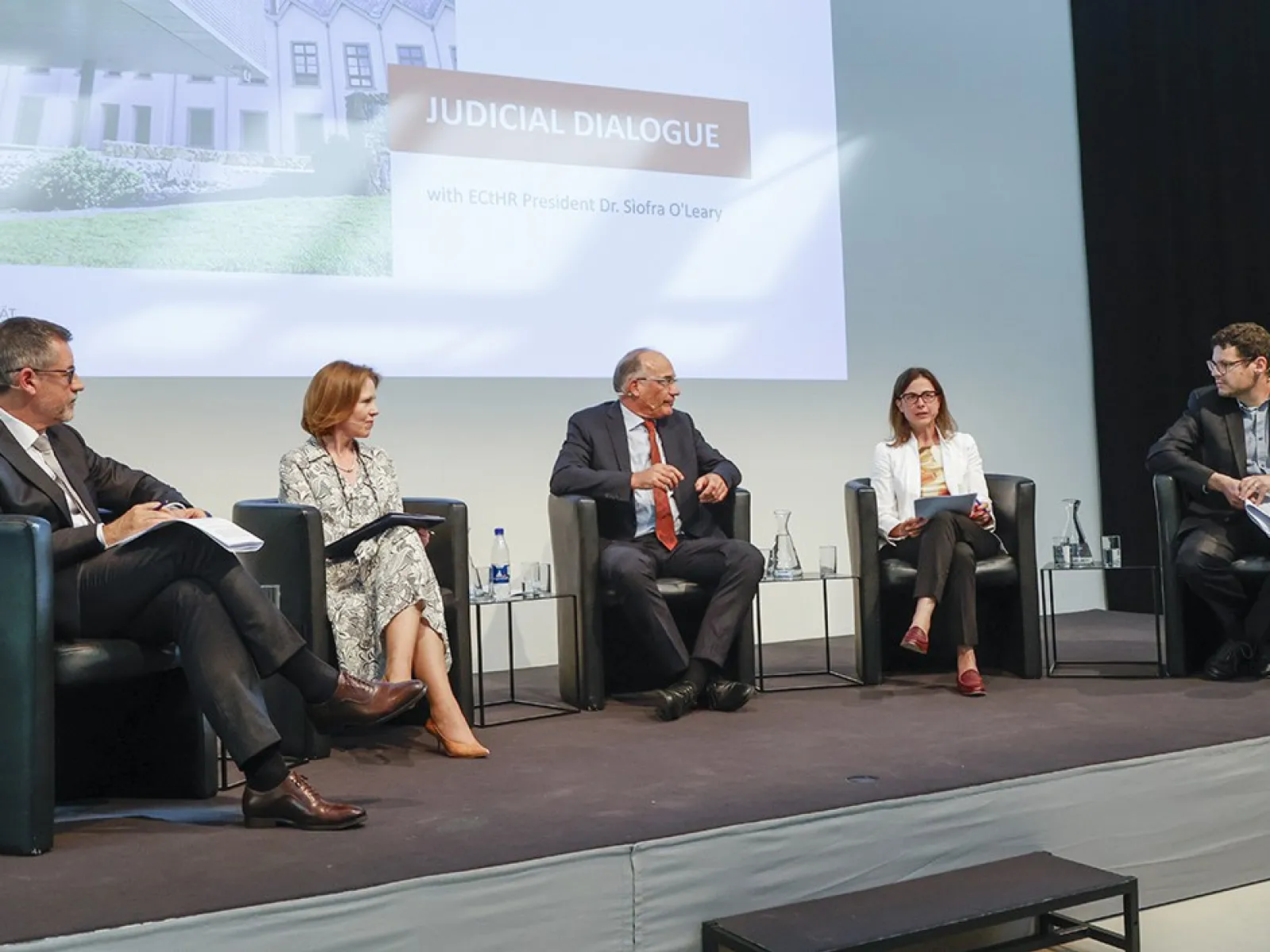 Podiumsdiskussion an der Universität Liechtenstein mit fünf Personen auf einer Bühne, die an einem juristischen Dialog teilnehmen. Im Hintergrund ist eine Präsentation mit dem Titel "Judicial Dialogue" zu sehen.
