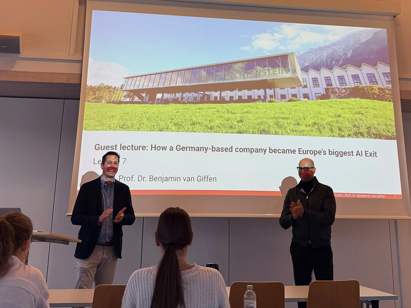 Zwei Männer stehen vor einer Präsentationsfolie in einem Vorlesungssaal der Universität Liechtenstein. Die Folie zeigt den Titel eines Gastvortrags über den Erfolg eines deutschen KI-Unternehmens. Studierende sitzen im Publikum.