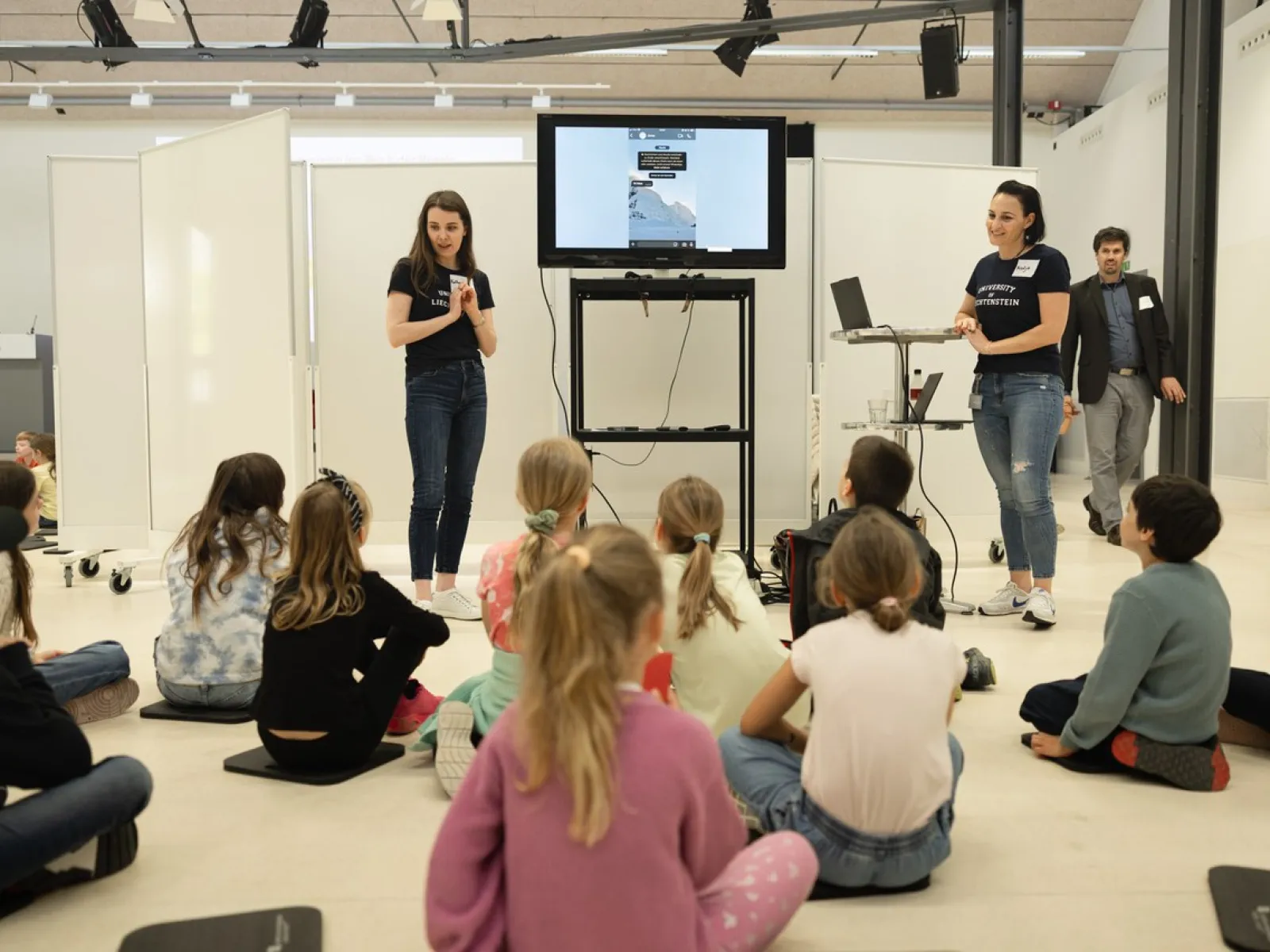 Eine Gruppe von Kindern sitzt auf dem Boden und hört zwei Frauen zu, die vor einem Bildschirm präsentieren. Die Frauen tragen dunkelblaue T-Shirts und Jeans. Im Hintergrund ist ein Mann zu sehen. Der Raum ist hell und modern gestaltet.