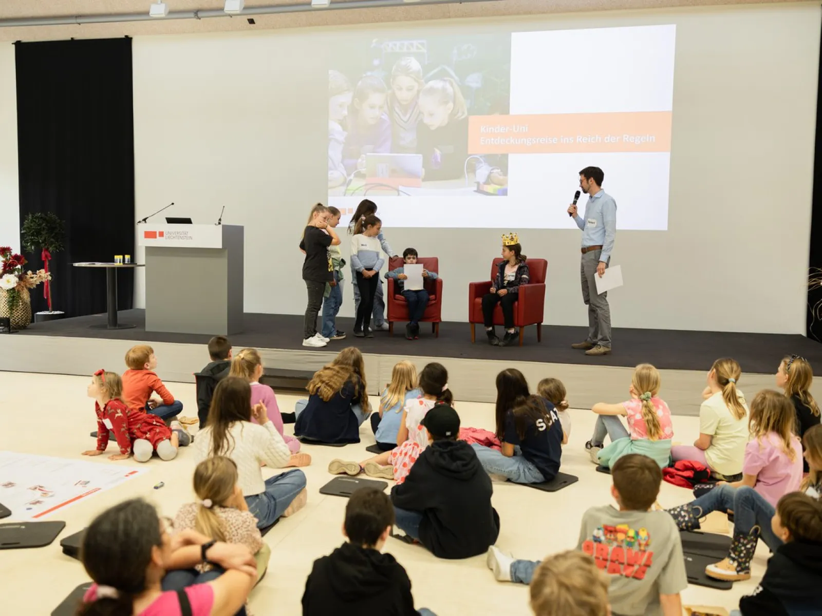 Ein Vortragssaal mit Kindern, die auf dem Boden sitzen und einem Vortragenden zuhören. Einige Kinder stehen auf der Bühne und nehmen an einer interaktiven Aktivität teil. Im Hintergrund ist eine Präsentation zu sehen.