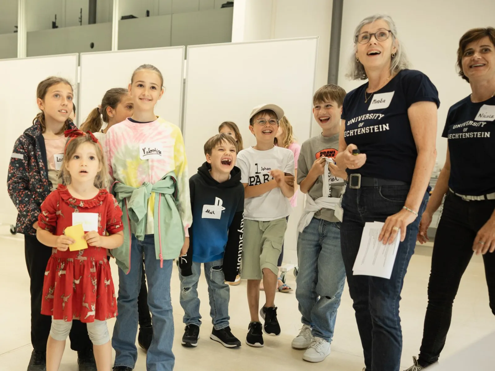 Eine Gruppe von Kindern und zwei Erwachsenen steht in einem hellen Raum. Die Kinder wirken fröhlich und interessiert. Die Erwachsenen tragen T-Shirts mit dem Logo der Universität Liechtenstein.