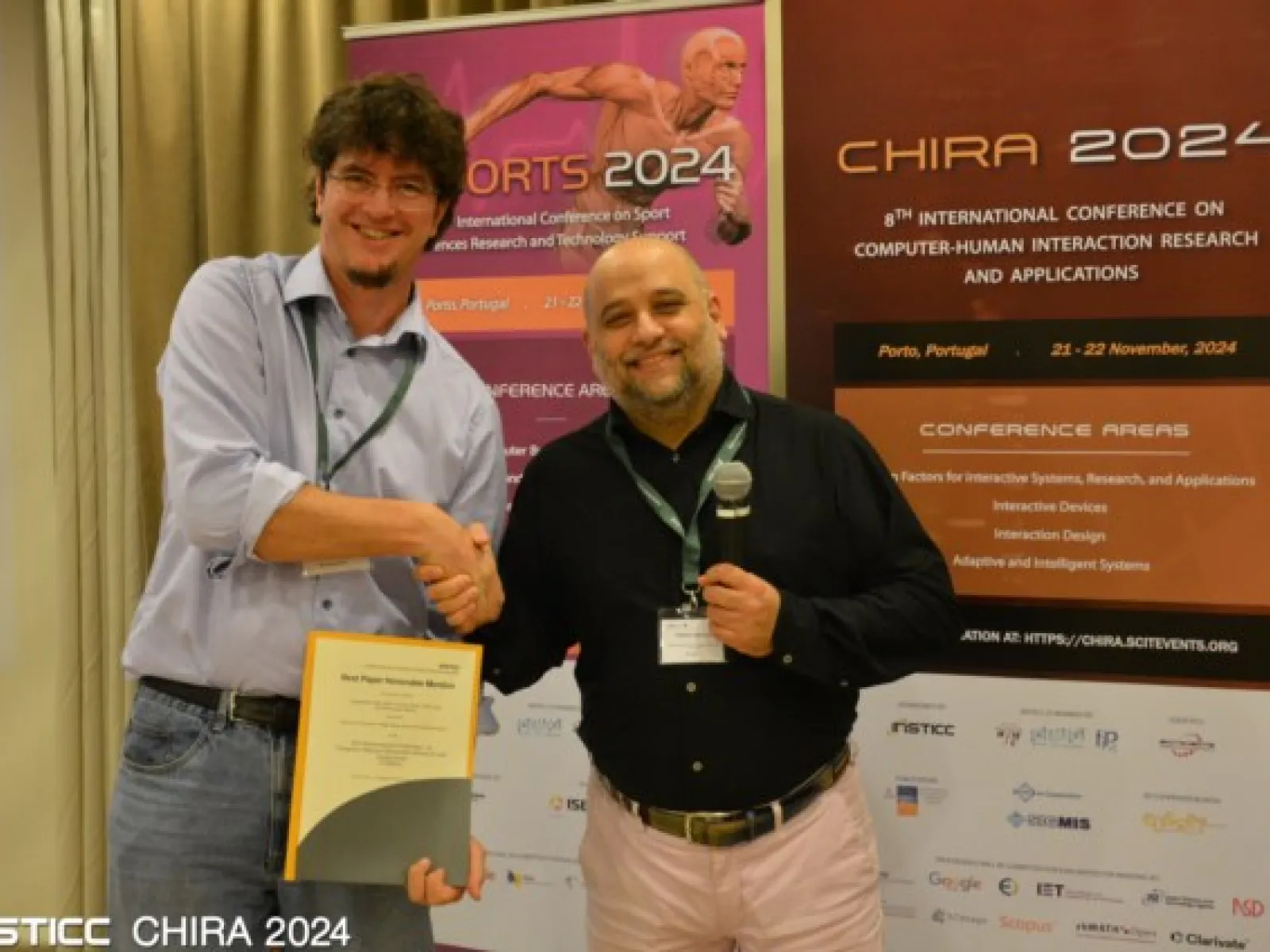 Zwei Männer stehen vor Bannern der CHIRA 2024 Konferenz. Einer hält ein Zertifikat, während der andere ein Mikrofon hält. Beide lächeln und schütteln sich die Hand. Im Hintergrund sind Details zur Konferenz in Porto, Portugal, sichtbar.