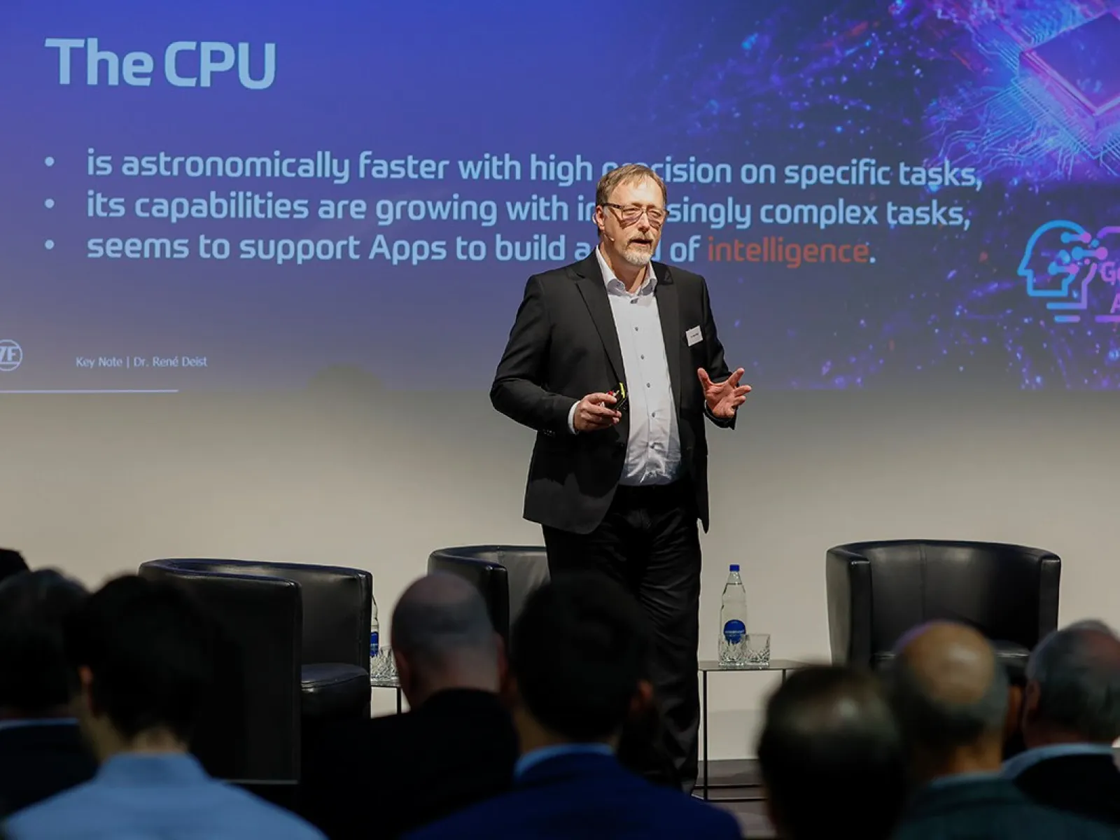 Ein Redner steht auf einer Bühne vor einem Publikum und präsentiert eine Folie über die Leistungsfähigkeit von CPUs bei einer Konferenz zur Künstlichen Intelligenz an der Universität Liechtenstein.