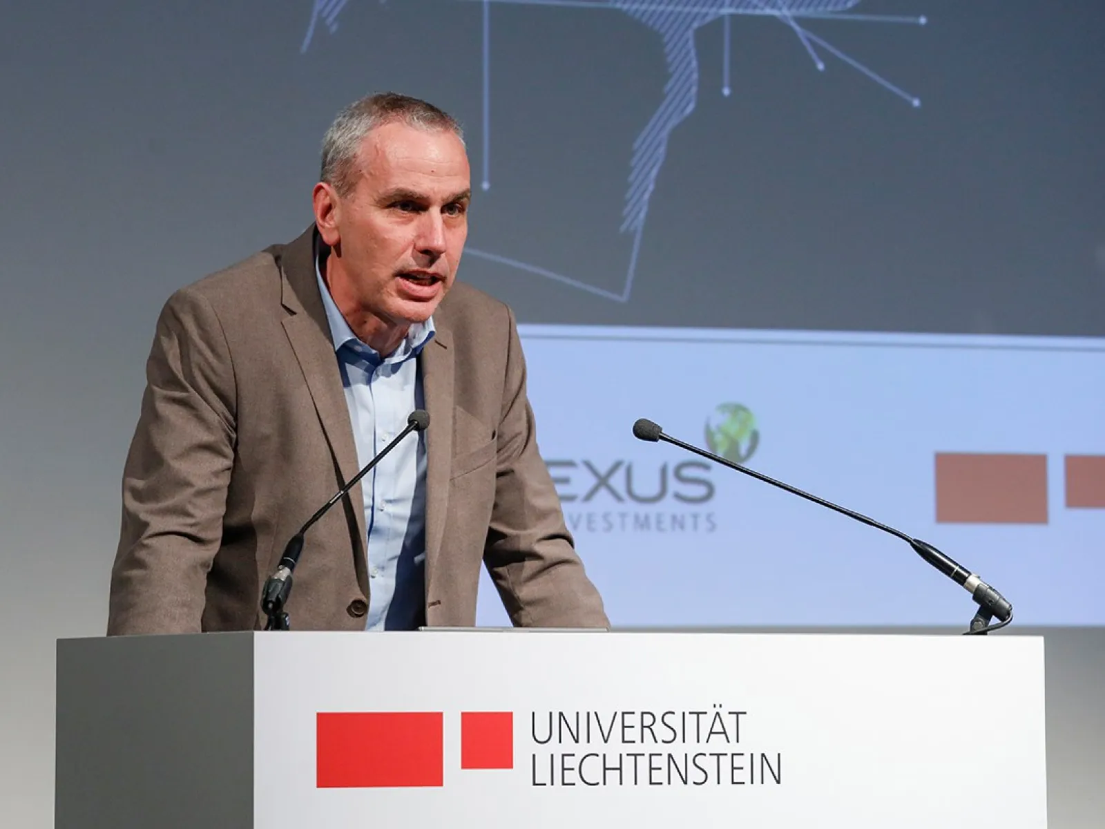 Ein Redner steht an einem Podium mit dem Logo der Universität Liechtenstein. Im Hintergrund sind Logos von PLEXUS Investments und der Universität Liechtenstein sichtbar.