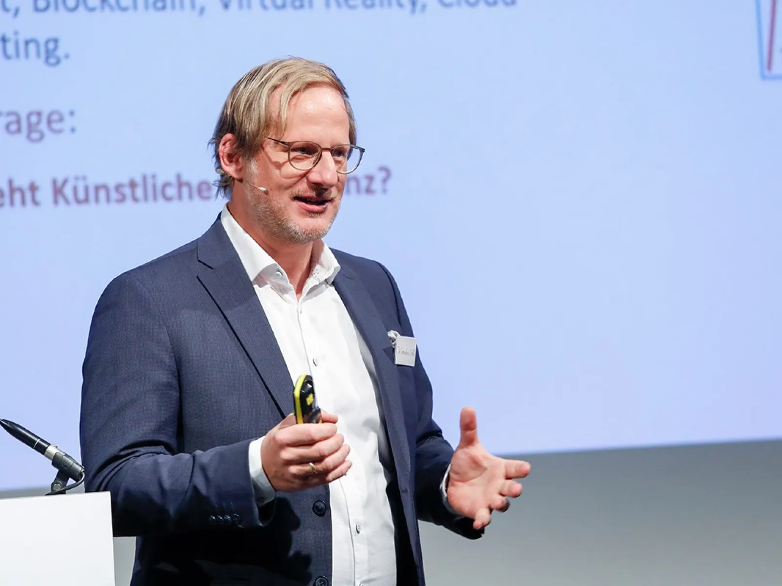 Ein Redner in einem Anzug hält einen Vortrag auf einer Konferenz über Künstliche Intelligenz an der Universität Liechtenstein. Im Hintergrund ist eine Präsentationsfolie zu sehen.