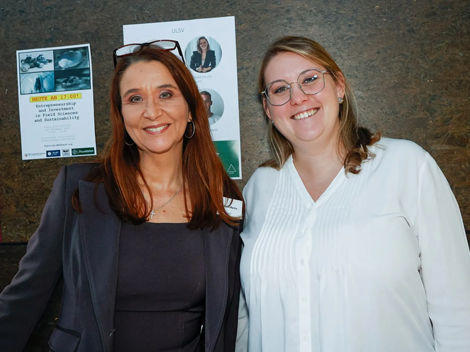 Zwei lächelnde Personen stehen vor einer Wand mit mehreren Postern. Eine Person trägt einen Blazer, die andere ein weißes Hemd.