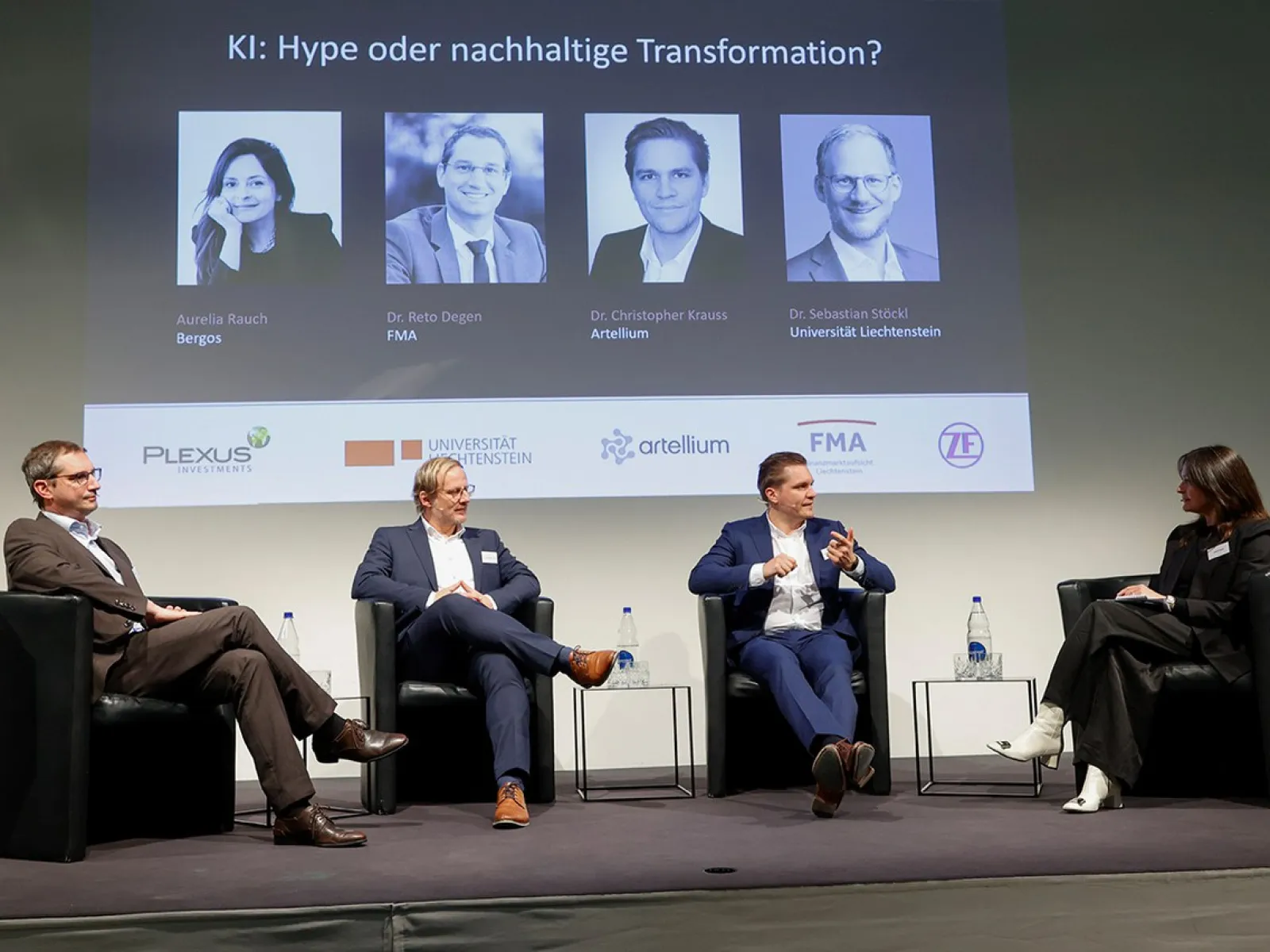 Eine Podiumsdiskussion an der Universität Liechtenstein über Künstliche Intelligenz mit vier Personen auf der Bühne. Im Hintergrund sind Porträts und Namen der Redner auf einer Leinwand zu sehen.