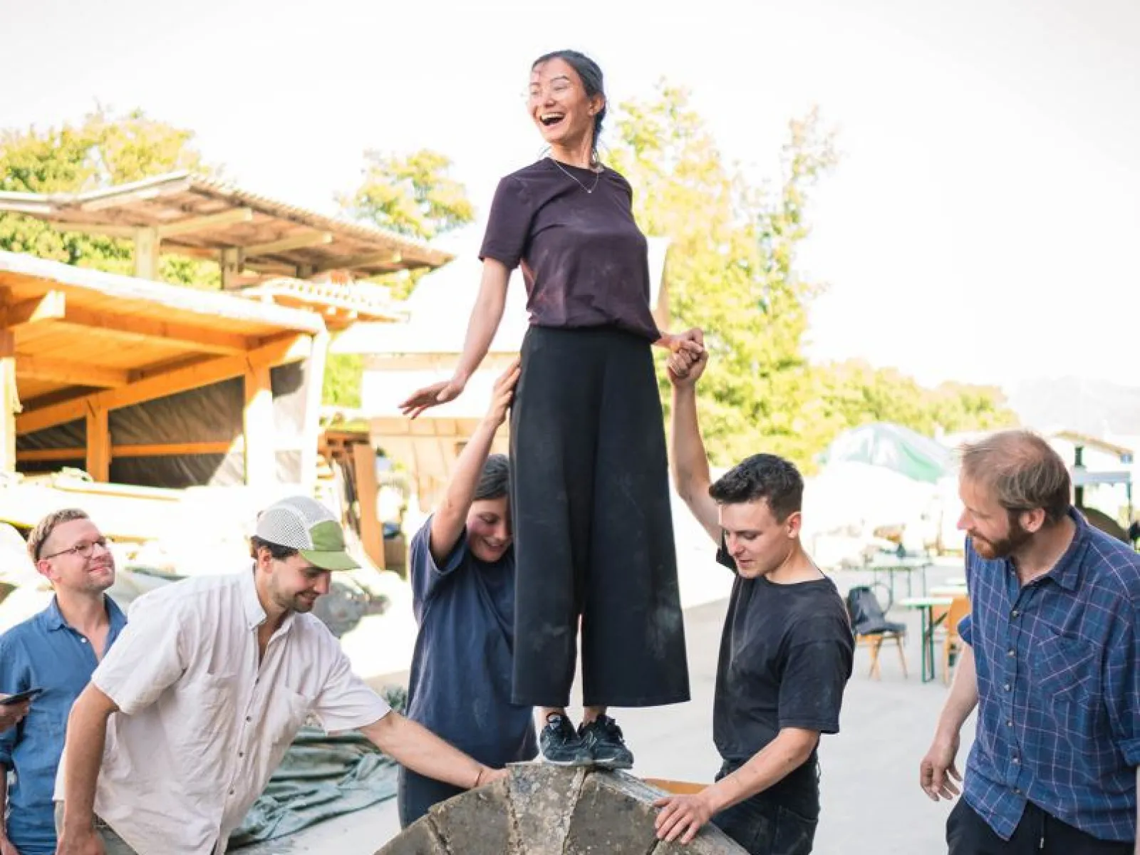 Ein Gruppe von Menschen baut einen selbsttragenden Lehmbogen im Freien. Eine Person steht oben auf dem Bogen, während andere sie stützen. Im Hintergrund sind Bäume und Gebäude zu sehen.
