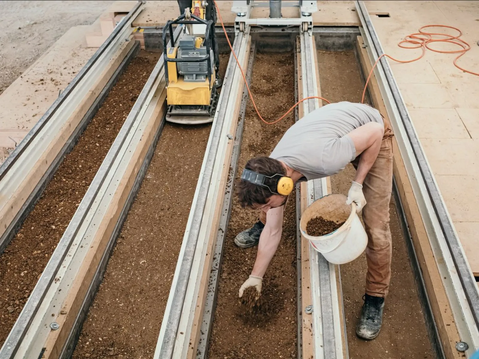 Eine Person arbeitet an einer Lehmbau-Konstruktion, indem sie Erde in eine Form füllt. Im Hintergrund ist eine Maschine zu sehen, die möglicherweise zur Verdichtung des Materials verwendet wird. Die Szene zeigt praktische Anwendung von Lehmbau-Techniken im EARTH HUB der Universität Liechtenstein.
