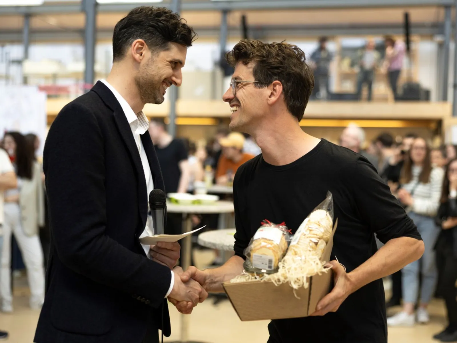Zwei Männer schütteln sich die Hand bei einer Veranstaltung in einem Innenraum. Einer hält ein Mikrofon, der andere ein Geschenkpaket. Im Hintergrund sind weitere Personen unscharf zu erkennen.