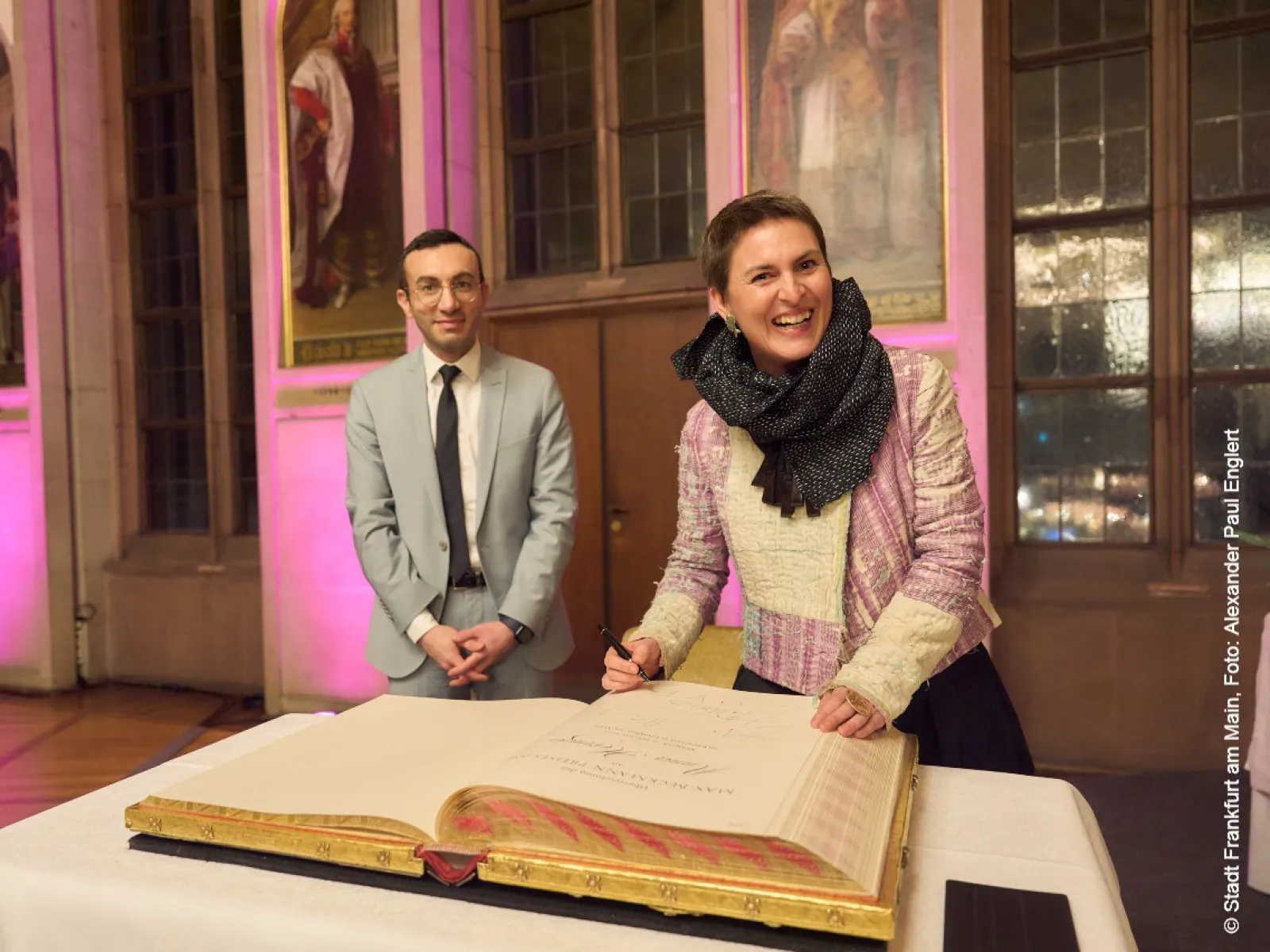 Eine Person in einem eleganten Outfit unterschreibt ein großes Buch auf einem Tisch, während eine andere Person daneben steht. Der Raum ist festlich beleuchtet und im Hintergrund sind Gemälde an der Wand zu sehen.