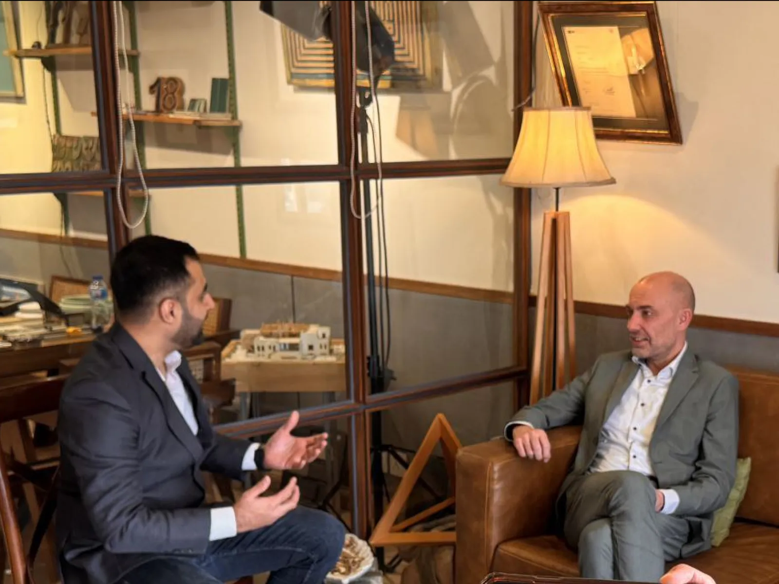 Zwei Männer in einem Gespräch in einem gemütlichen Raum mit Glaswänden und Holzmöbeln. Einer sitzt auf einem Ledersessel, der andere gestikuliert während des Gesprächs. Im Hintergrund sind Bücher und Dekorationsgegenstände zu sehen.