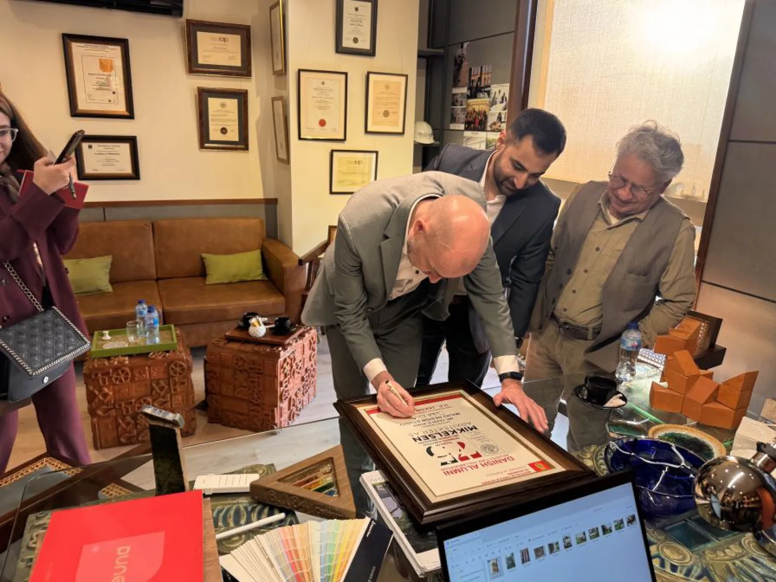 Ein Mann in einem Anzug unterschreibt eine Auszeichnung in einem Büro, während zwei weitere Männer zuschauen. Im Hintergrund sind gerahmte Zertifikate an der Wand zu sehen. Auf dem Tisch liegen Farbmuster und ein Laptop.