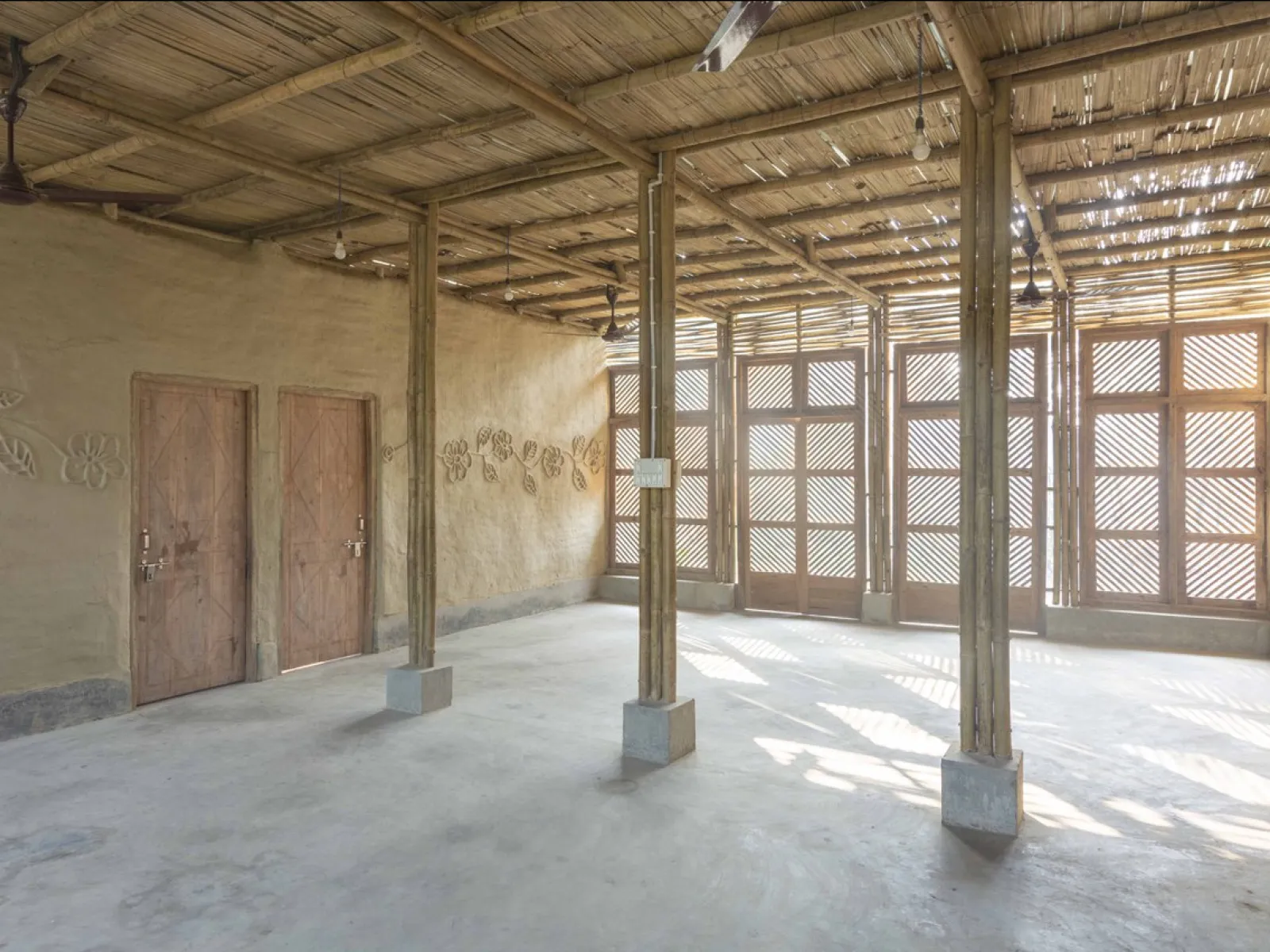 Alt-Text: Innenansicht eines nachhaltig gebauten Raumes mit Bambusstrukturen, Lehmwänden und Holztüren. Sonnenlicht fällt durch ein Gittermuster an den Fenstern und erzeugt ein warmes Ambiente.