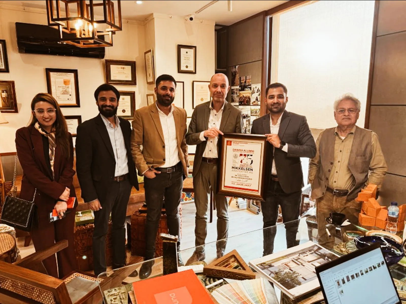 Eine Gruppe von sechs Personen steht in einem Büro, eine Person hält eine gerahmte Auszeichnung. Im Hintergrund sind Zertifikate und Auszeichnungen an der Wand zu sehen.