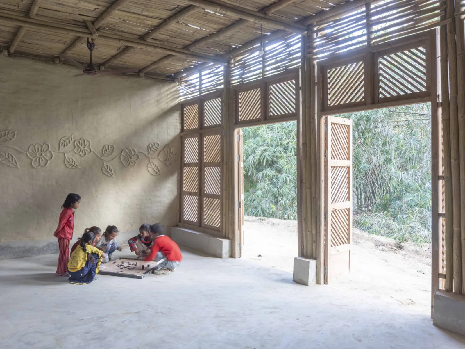 Einige Kinder spielen auf dem Boden eines hellen, nachhaltig gebauten Raumes mit großen Holztüren und dekorativen Wänden.