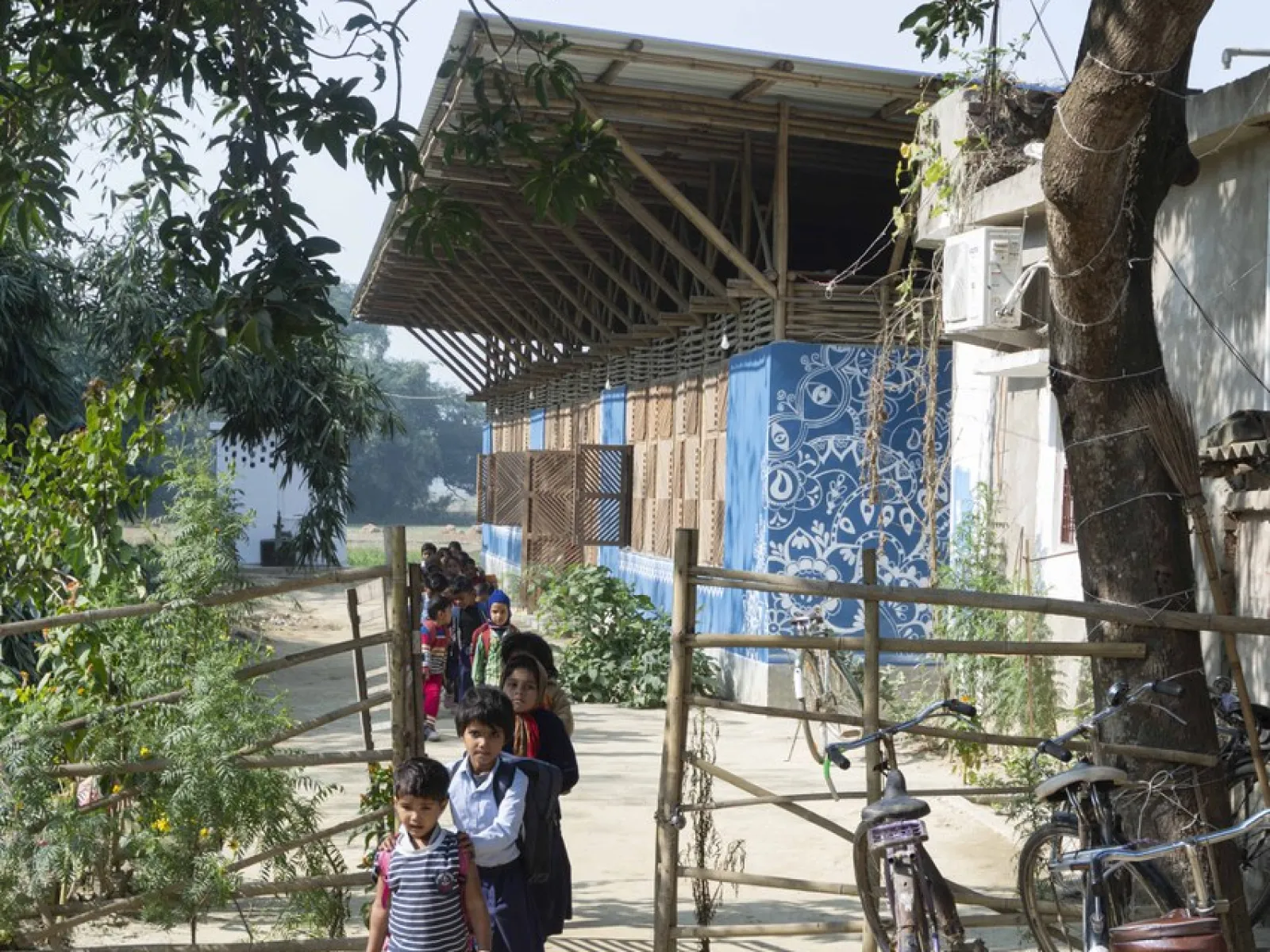 Eine Gruppe von Kindern geht einen Weg entlang, der zu einem nachhaltig gebauten Gebäude mit blauen Wänden und einem großen Dach führt. Im Vordergrund sind Bäume und ein Zaun zu sehen. Die Szene vermittelt eine ländliche, sonnige Atmosphäre.