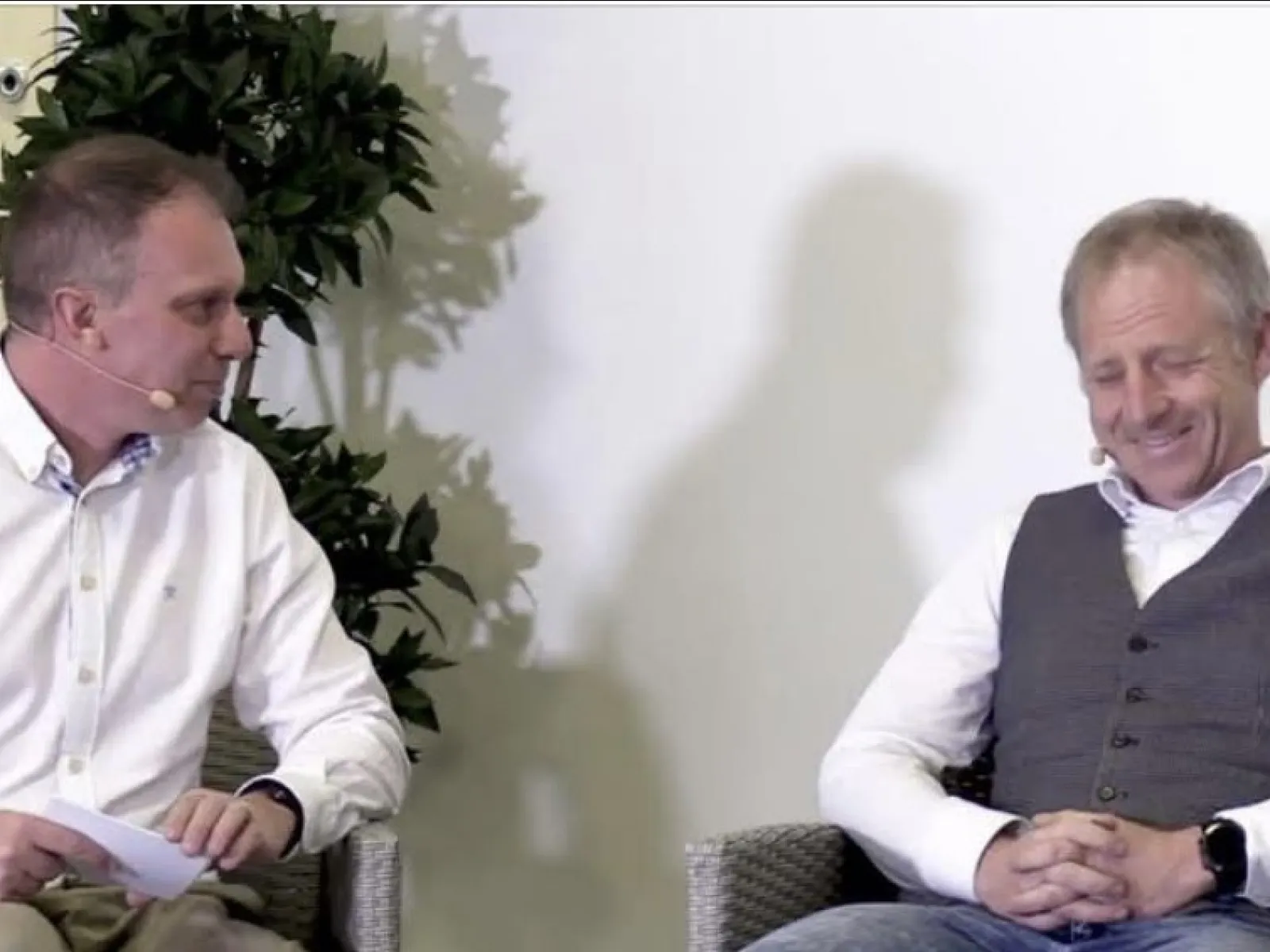 Zwei Männer sitzen auf einer Bühne und unterhalten sich. Beide tragen Mikrofone. Links sitzt ein Mann in einem weißen Hemd, rechts ein Mann in einem grauen Pullunder. Im Hintergrund ist eine Pflanze zu sehen.