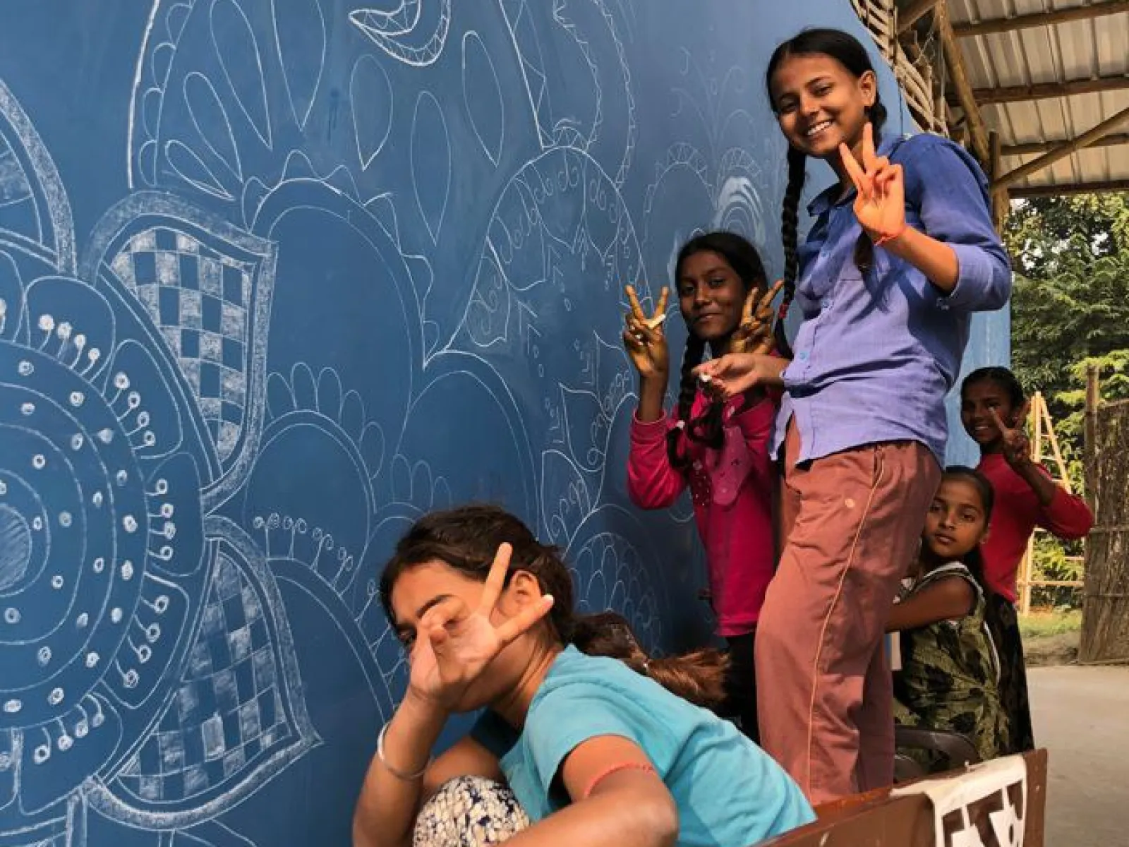 Kinder posieren fröhlich vor einer blau bemalten Wand mit kunstvollen weißen Mustern. Sie zeigen mit den Fingern das Friedenszeichen. Die Szene vermittelt Freude und Gemeinschaftsgefühl.