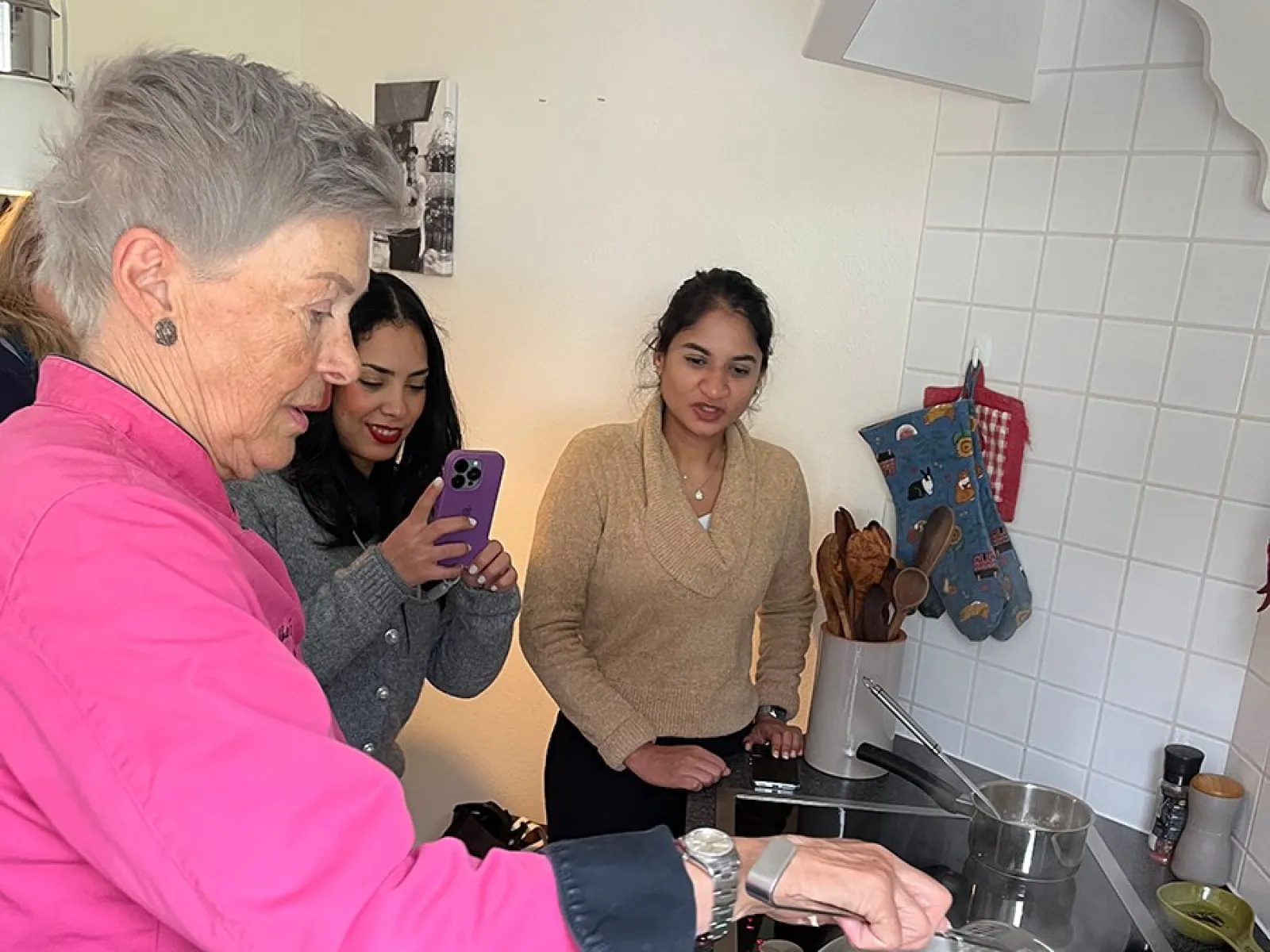 Eine Gruppe von Menschen steht in einer Küche. Eine Person in einem pinken Oberteil kocht an einem Herd, während zwei andere Personen zuschauen und eine von ihnen mit einem Smartphone fotografiert. Auf dem Herd steht ein Topf mit kochendem Wasser.