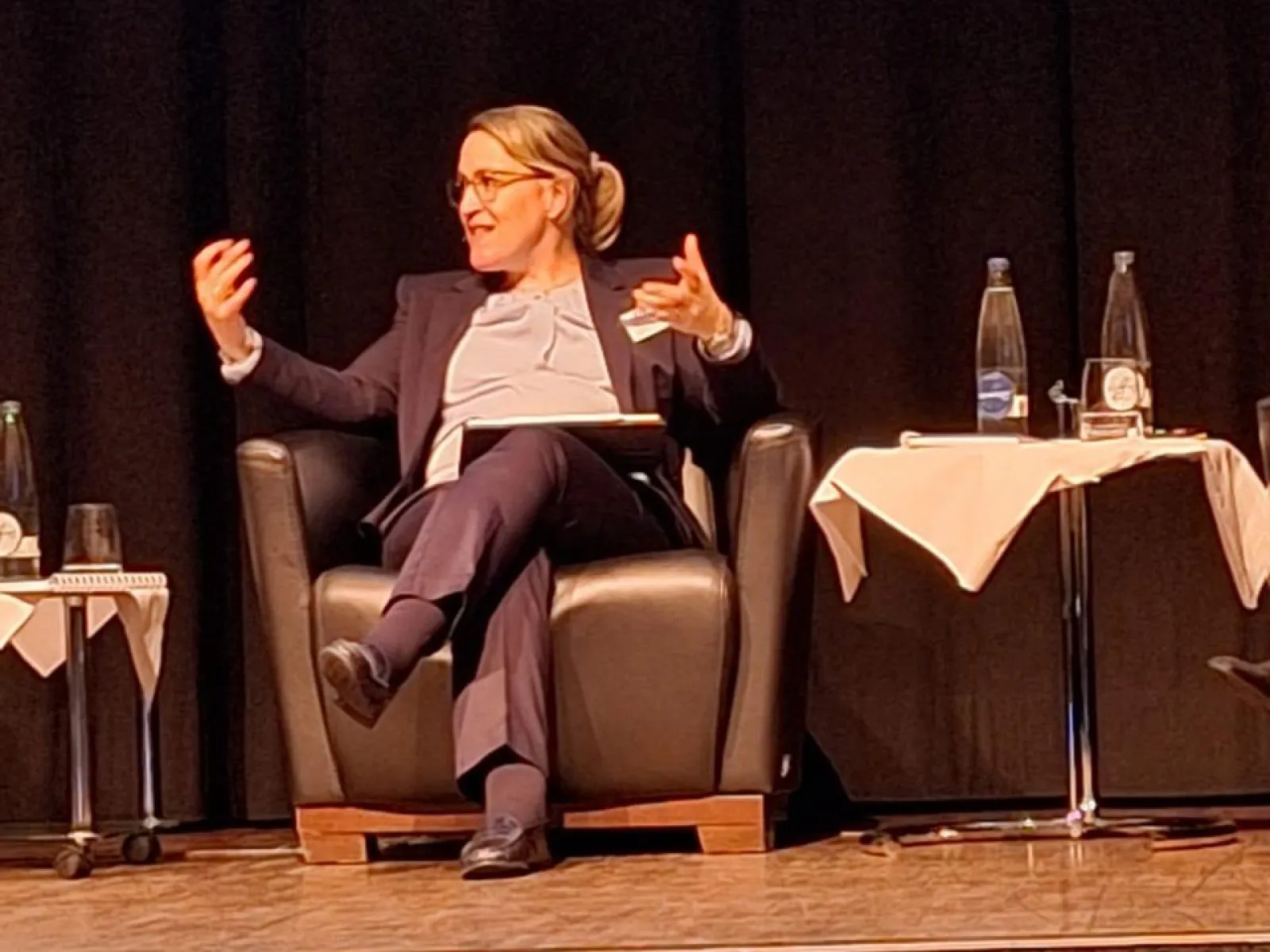 Eine Person sitzt auf einem Podium in einem Sessel und gestikuliert während einer Diskussion. Neben ihr stehen Tische mit Wasserflaschen und Gläsern. Die Szene findet in einem Konferenzsaal statt.