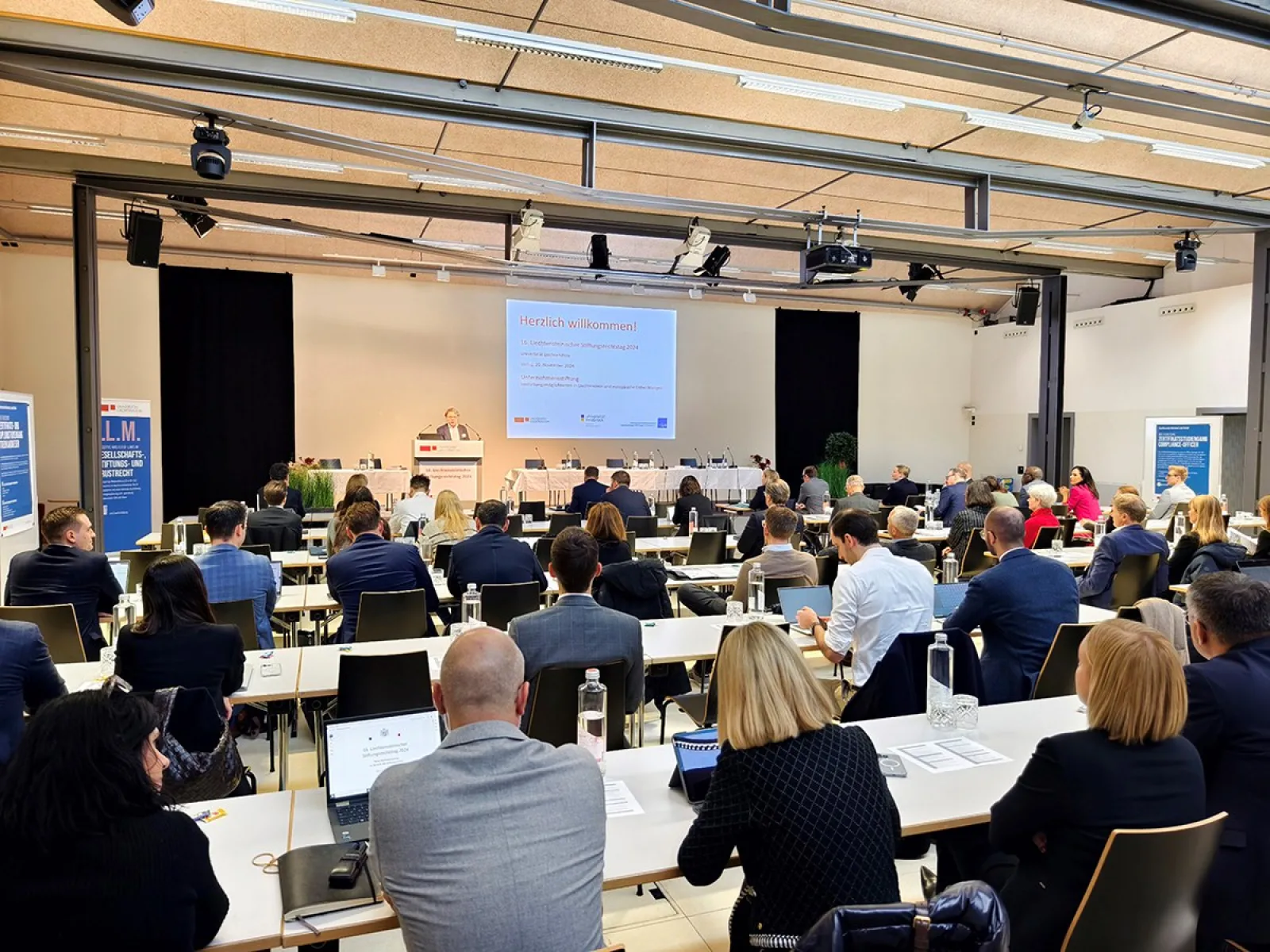 Ein Auditorium voller Menschen sitzt an Tischen und hört einem Vortrag zu. Auf einer Leinwand vorne sind Präsentationsfolien zu sehen. Die Veranstaltung findet in einem modernen, gut beleuchteten Raum statt.