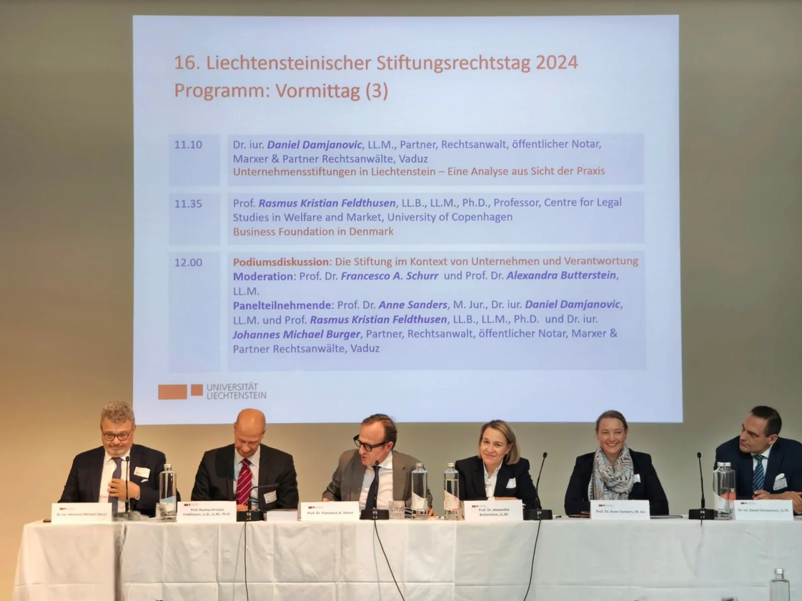 Podiumsdiskussion beim 16. Liechtensteinischen Stiftungsrechtstag 2024 an der Universität Liechtenstein. Fünf Expertinnen und Experten sitzen an einem Tisch vor einer Leinwand mit dem Tagesprogramm.