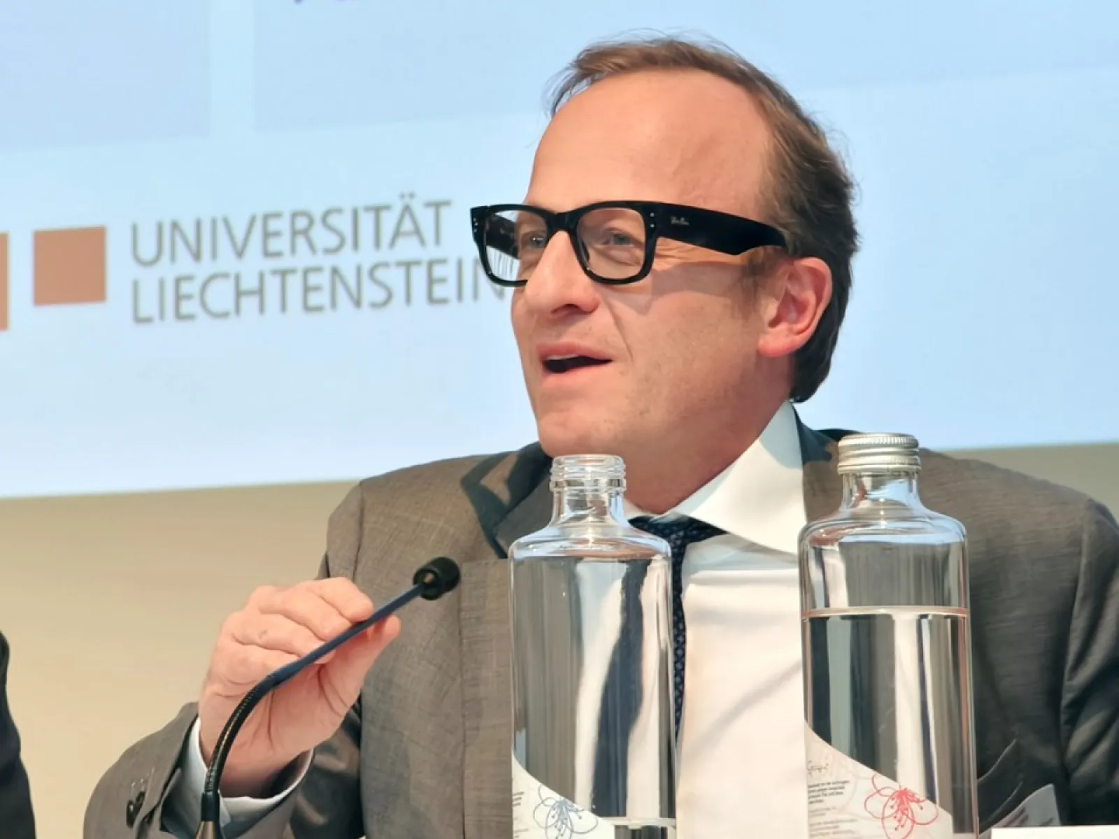 Ein Mann in Anzug und Brille spricht in ein Mikrofon auf einer Konferenz im Auditorium der Universität Liechtenstein. Vor ihm stehen Wasserflaschen. Im Hintergrund ist das Logo der Universität zu sehen.