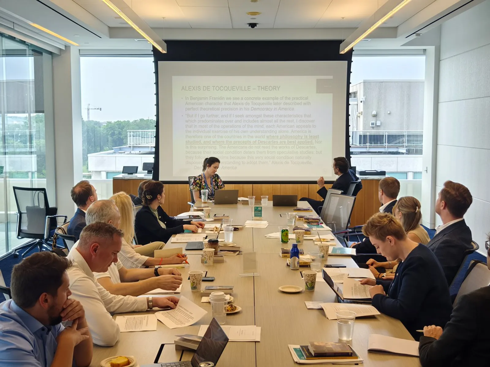 Ein Konferenzraum mit mehreren Personen, die an einem langen Tisch sitzen und einer Präsentation folgen. Im Hintergrund ist eine große Leinwand mit projiziertem Text zu sehen. Die Teilnehmer sind in eine Diskussion vertieft.