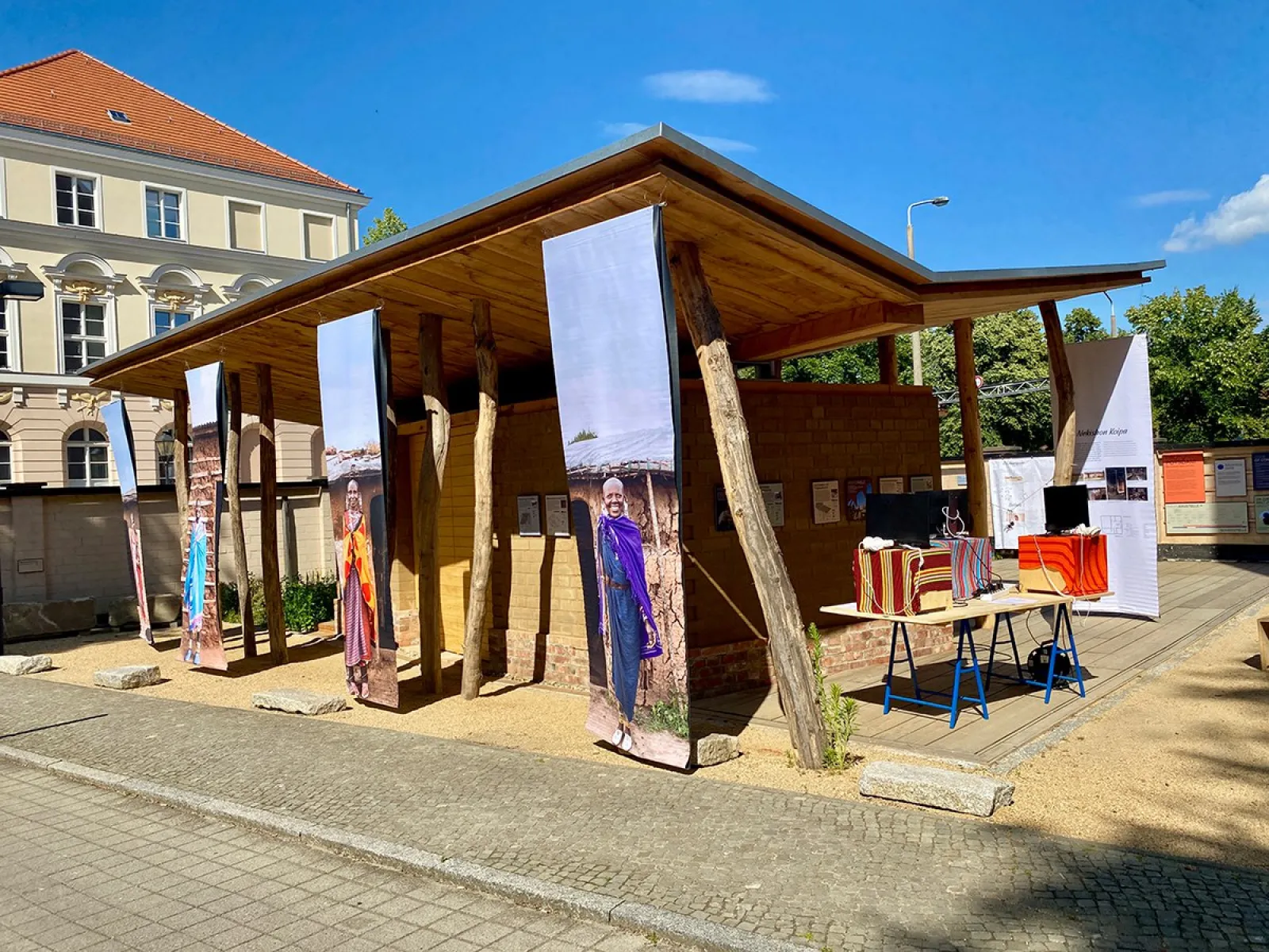 Ein hölzerner Pavillon mit großen, vertikalen Bannern, die Maasai-Frauen in traditioneller Kleidung zeigen. Im Vordergrund stehen Tische mit bunten Stoffen und Modellen. Der Pavillon ist Teil einer Ausstellung über Maasai-Baumeisterinnen und nachhaltige Architektur.