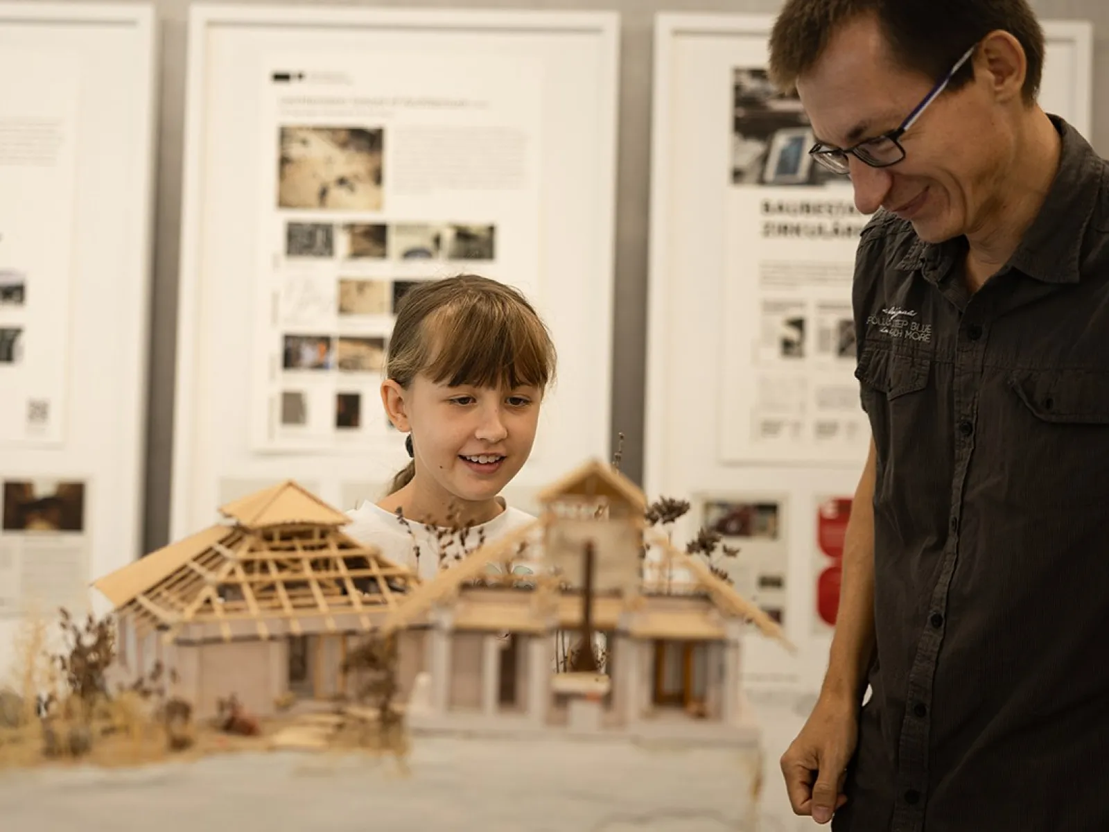 Ein Kind und ein Erwachsener betrachten ein detailliertes Architekturmodell eines Gebäudes. Im Hintergrund sind Informationsplakate zu sehen.