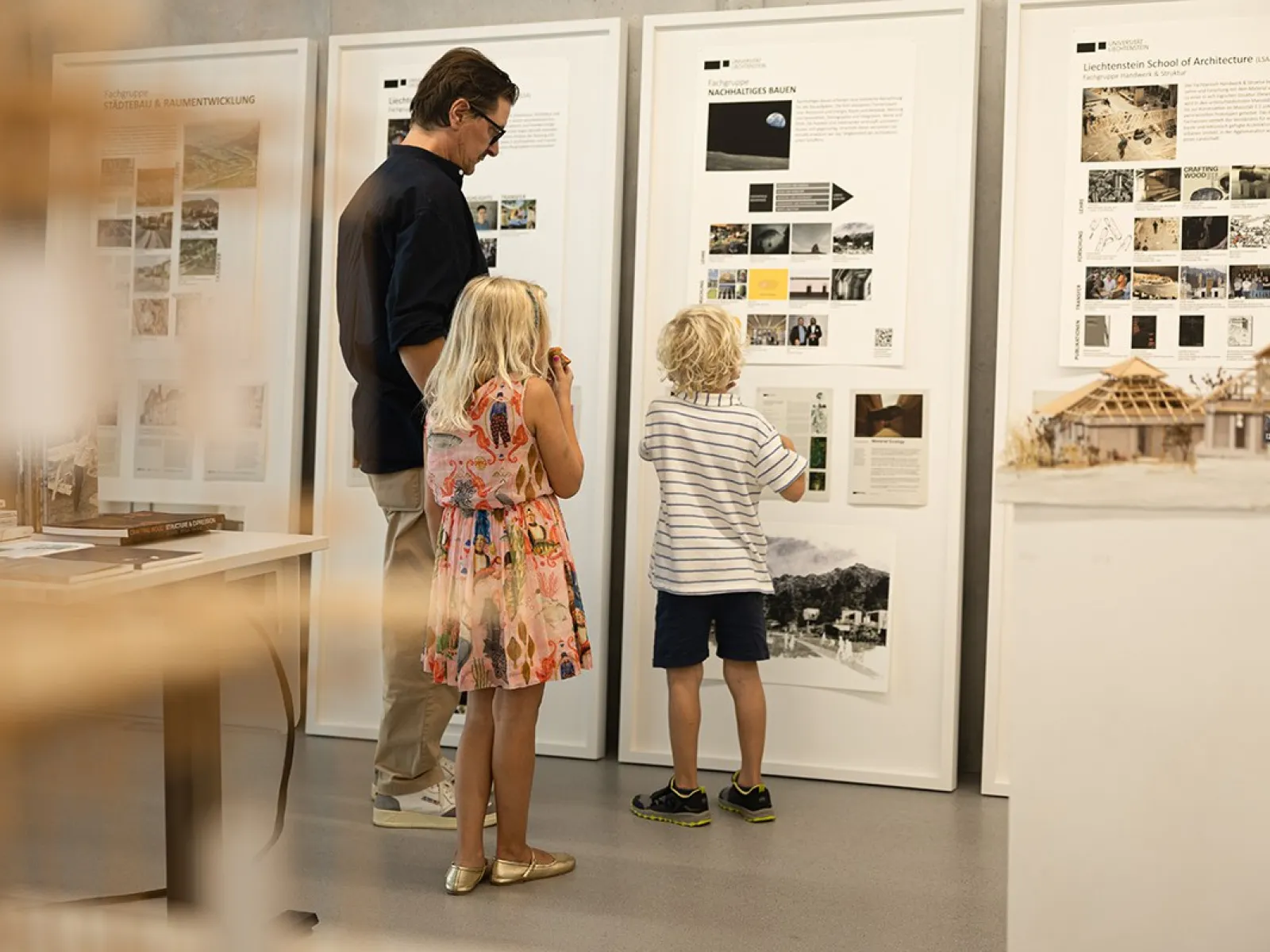 Ein Erwachsener und zwei Kinder betrachten Informationswände mit Architekturprojekten in einem Ausstellungsraum der Universität Liechtenstein.