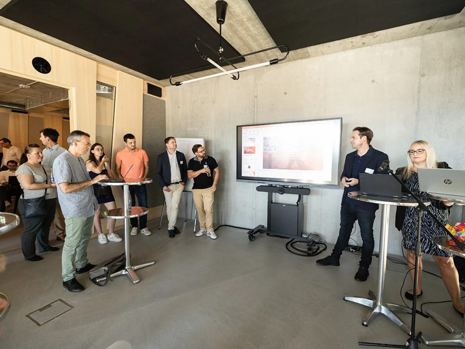 Eine Gruppe von Menschen steht in einem modernen Raum mit Betonwänden. Im Vordergrund sind Stehtische und ein großer Bildschirm an der Wand, auf dem eine Präsentation gezeigt wird. Eine Person spricht zu den Anwesenden, während andere aufmerksam zuhören. Der Raum wirkt hell und einladend.