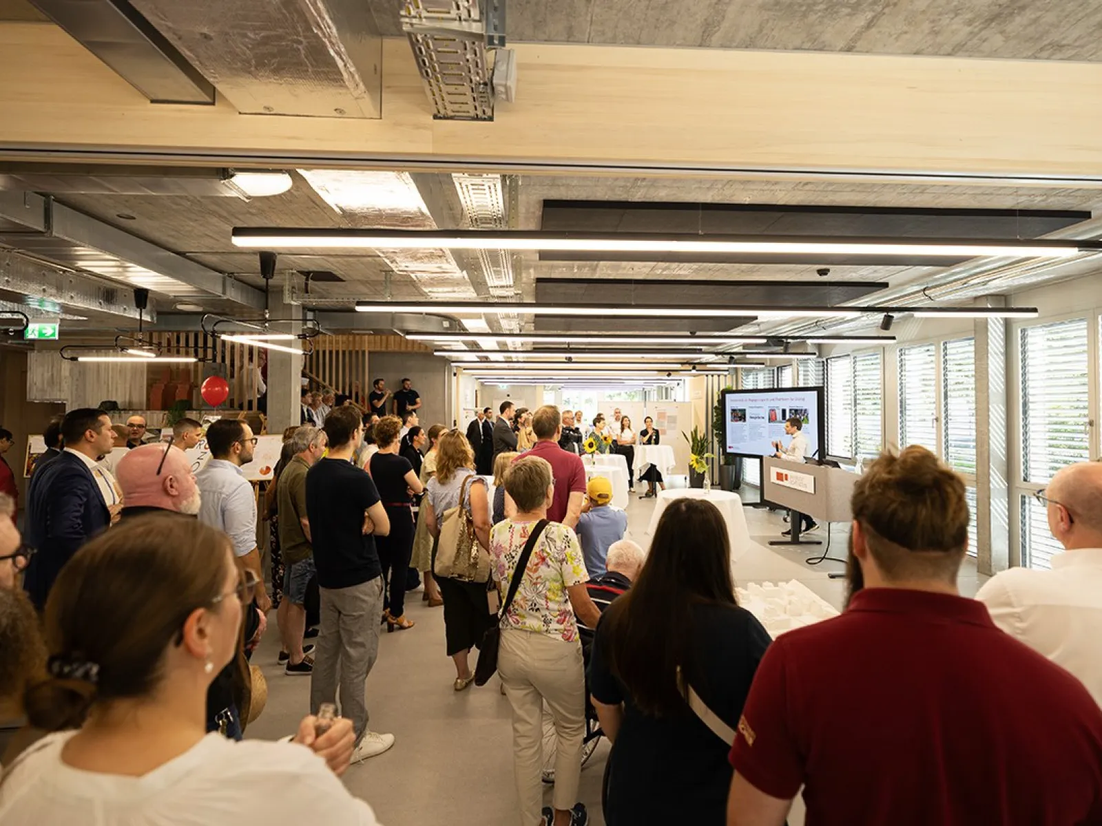 Besuchergruppe beim Tag der offenen Tür an der Universität Liechtenstein, neuer Standort Ebaholz. Menschen stehen in einem modernen Raum und hören einer Präsentation zu. Im Hintergrund sind große Fenster und eine Leinwand zu sehen.