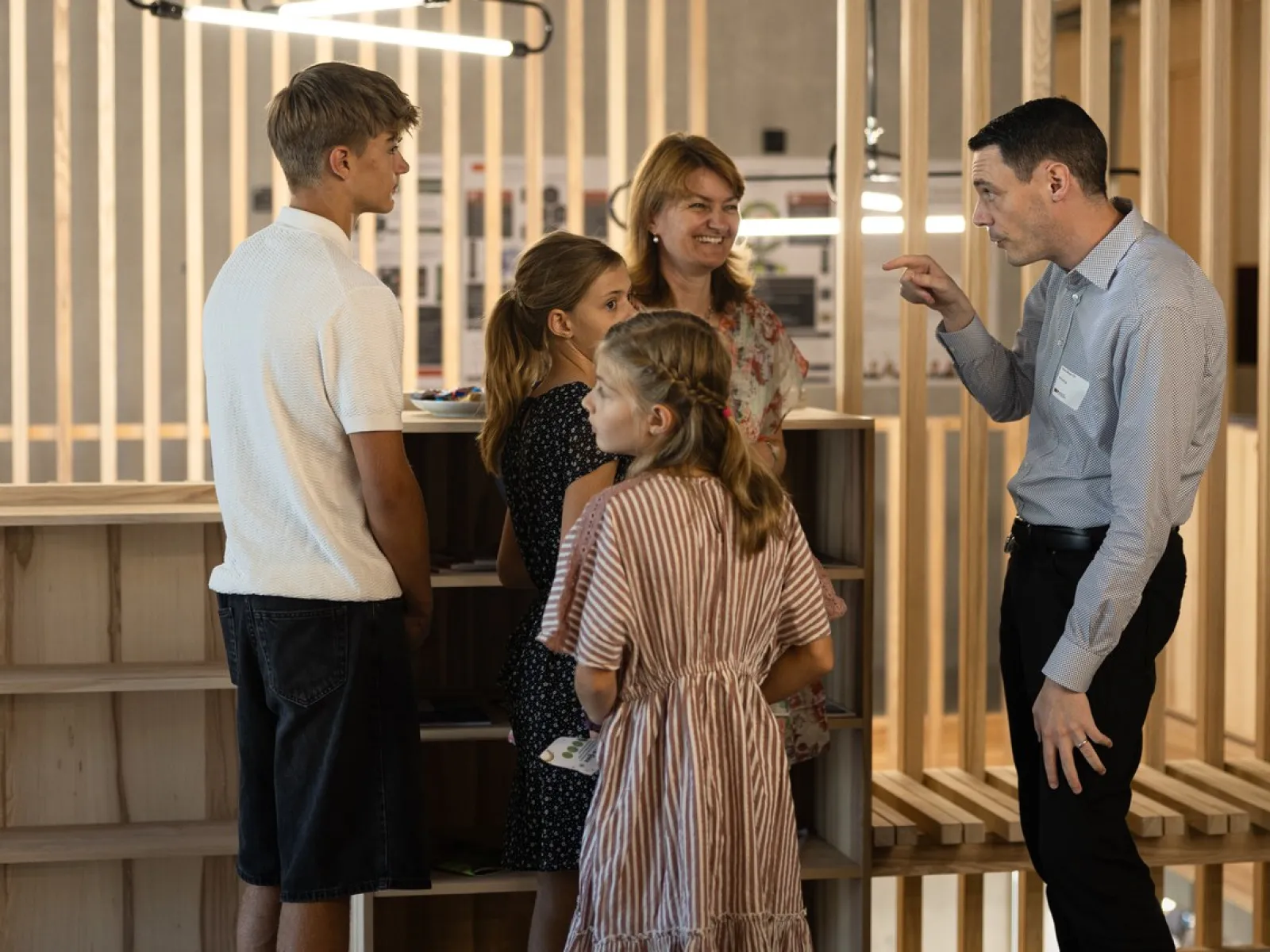 Eine Gruppe von Menschen steht in einem modernen Raum mit Holzelementen. Ein Mann spricht gestikulierend mit einer Frau und drei Kindern. Die Szene vermittelt eine freundliche und einladende Atmosphäre während des Tags der offenen Tür an der Universität Liechtenstein.