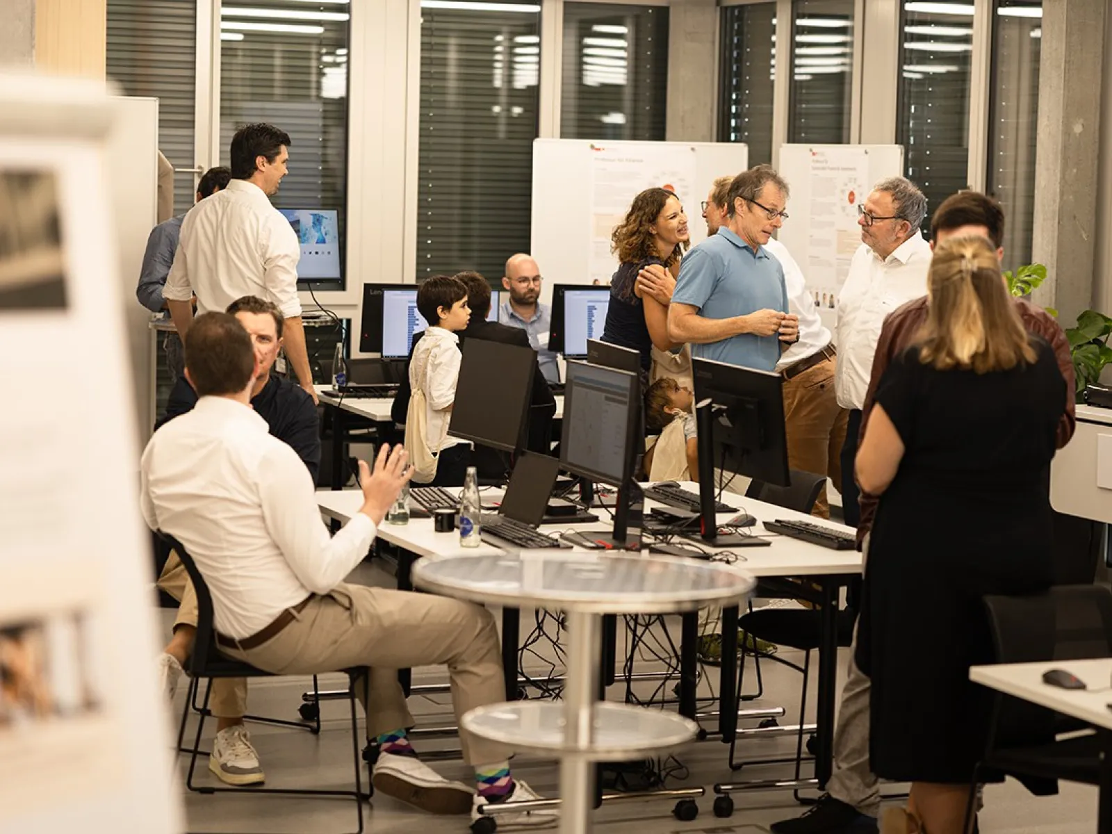 Eine Gruppe von Menschen unterhält sich in einem modernen Raum mit Computern und Informationspostern. Einige sitzen, während andere stehen und diskutieren. Die Atmosphäre wirkt lebhaft und interessiert, passend zu einem Tag der offenen Tür an der Universität.