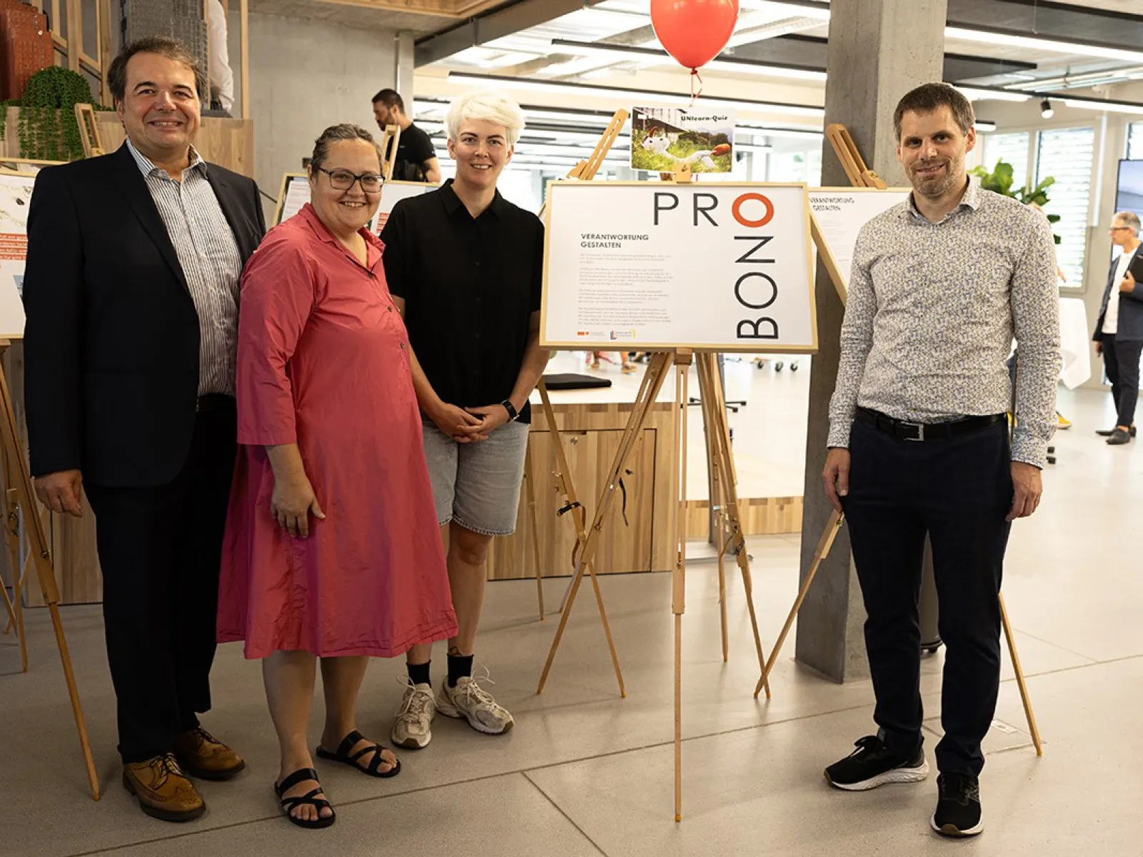 Vier Personen stehen in einem modernen Raum neben einer Ausstellungstafel mit der Aufschrift "Pro Bono". Im Hintergrund sind weitere Ausstellungstafeln und ein roter Luftballon zu sehen. Die Szene wirkt freundlich und einladend.