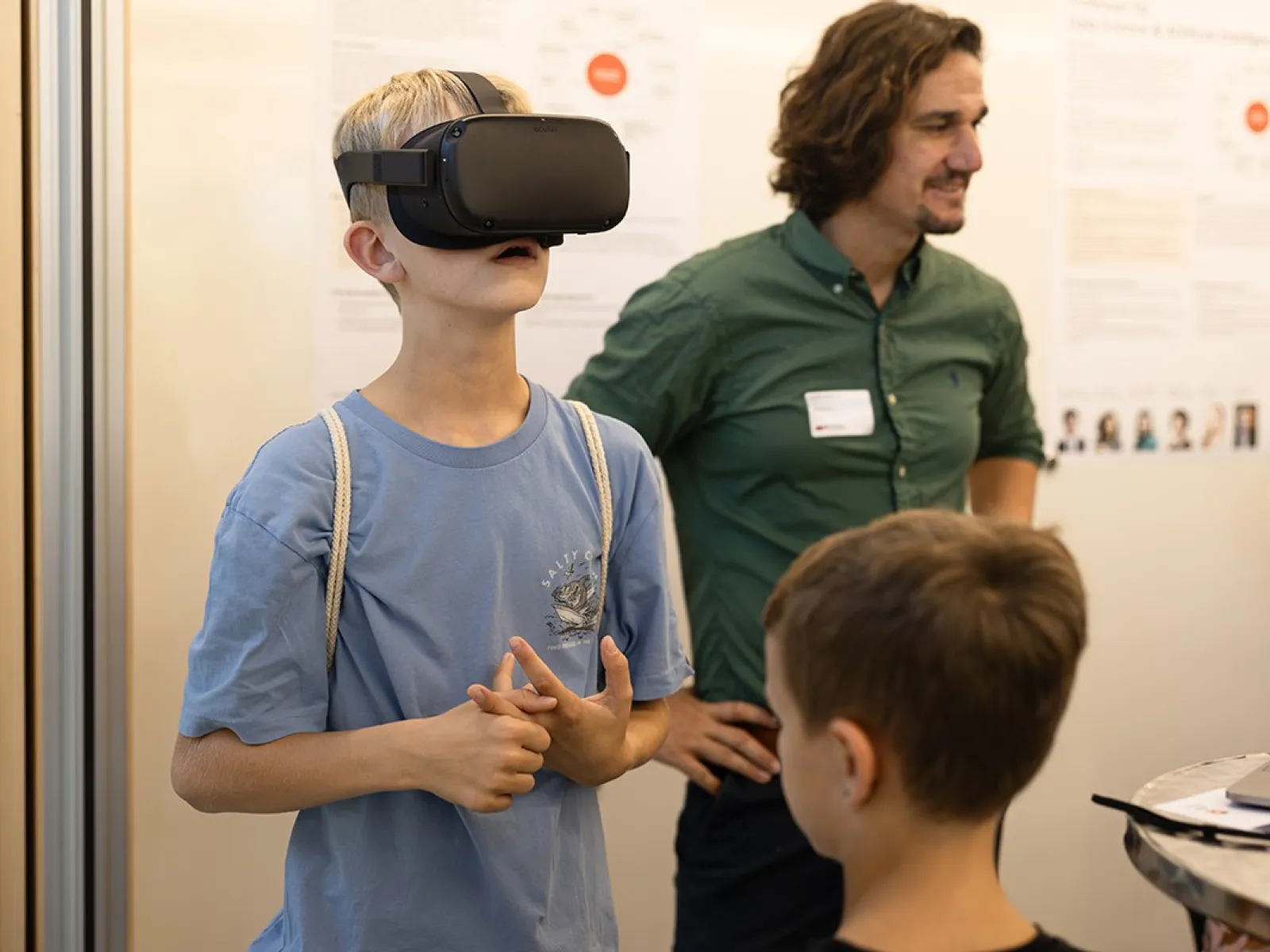 Ein Junge trägt eine VR-Brille und erkundet virtuelle Inhalte. Im Hintergrund steht ein Mann, der lächelnd zusieht. Weitere Personen sind teilweise sichtbar. Die Szene spielt in einem Raum mit Informationspostern an der Wand.