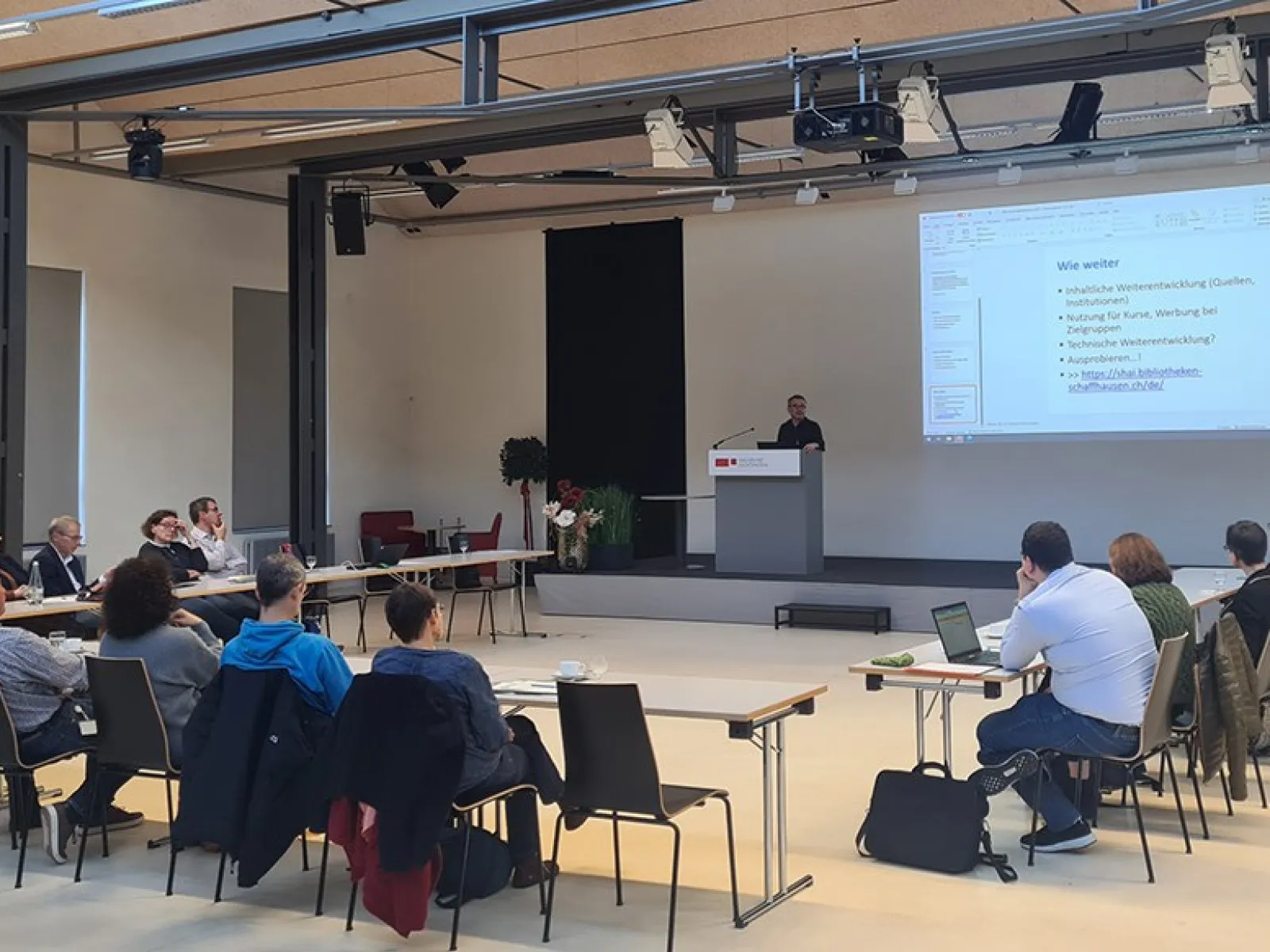 Eine Gruppe von etwa 20 Personen sitzt in einem Auditorium der Universität Liechtenstein, während ein Vortragender an einem Rednerpult spricht. Auf einer großen Leinwand ist eine Präsentation mit dem Titel "Wie weiter" zu sehen.