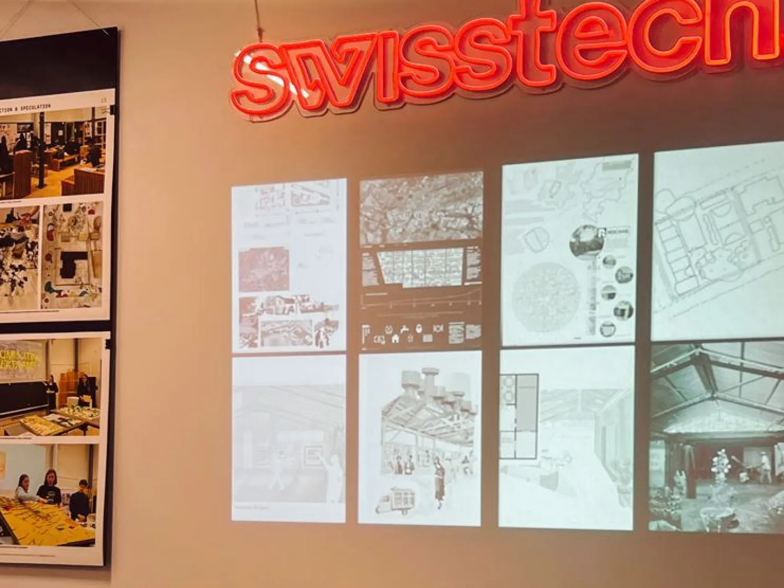 Ein Ausstellungstisch mit offenen Büchern steht vor einer Wand, auf der Architekturzeichnungen und Pläne projiziert sind. Links sind Fotos von Designprojekten zu sehen. Oben leuchtet ein "swisstech" Schild in roter Schrift.