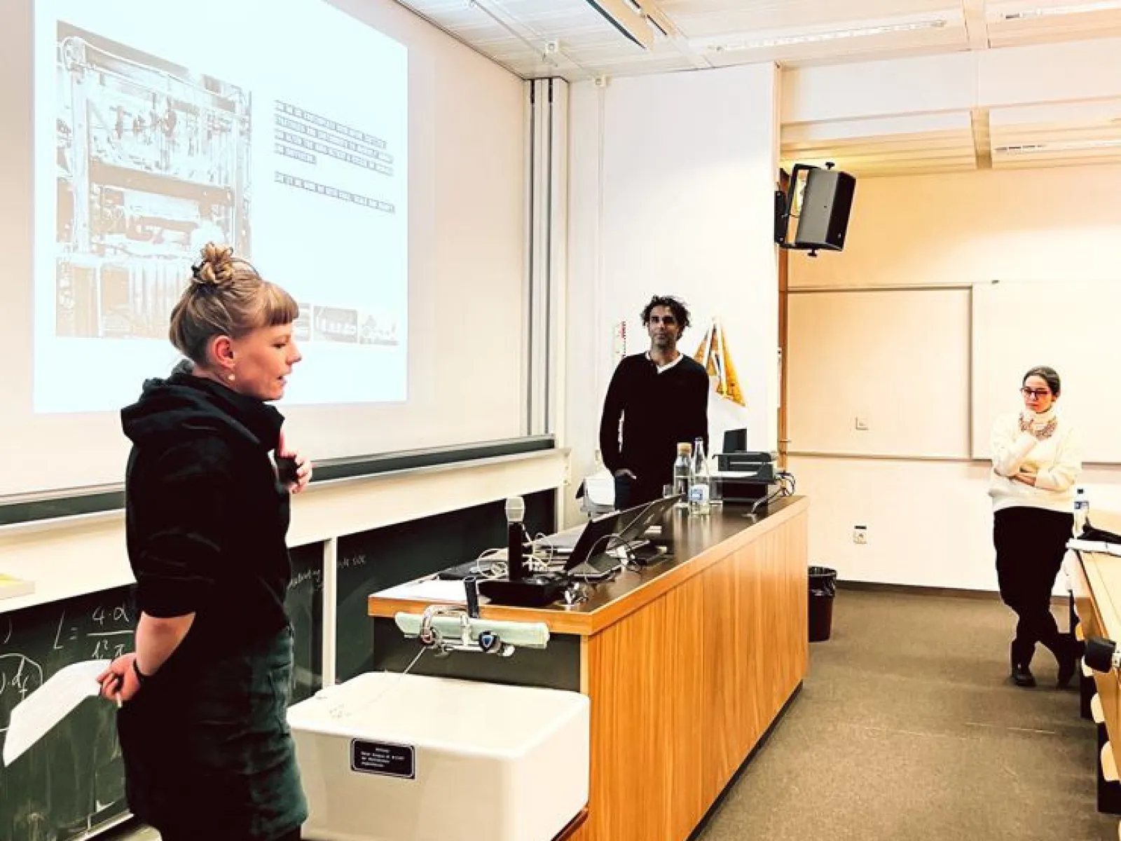 Eine Präsentation in einem Universitätsseminarraum: Drei Personen stehen vor einer Leinwand, auf der eine Folie projiziert wird. Eine Person spricht, während die anderen zuhören. Der Raum ist mit Tischen und einer Tafel ausgestattet.